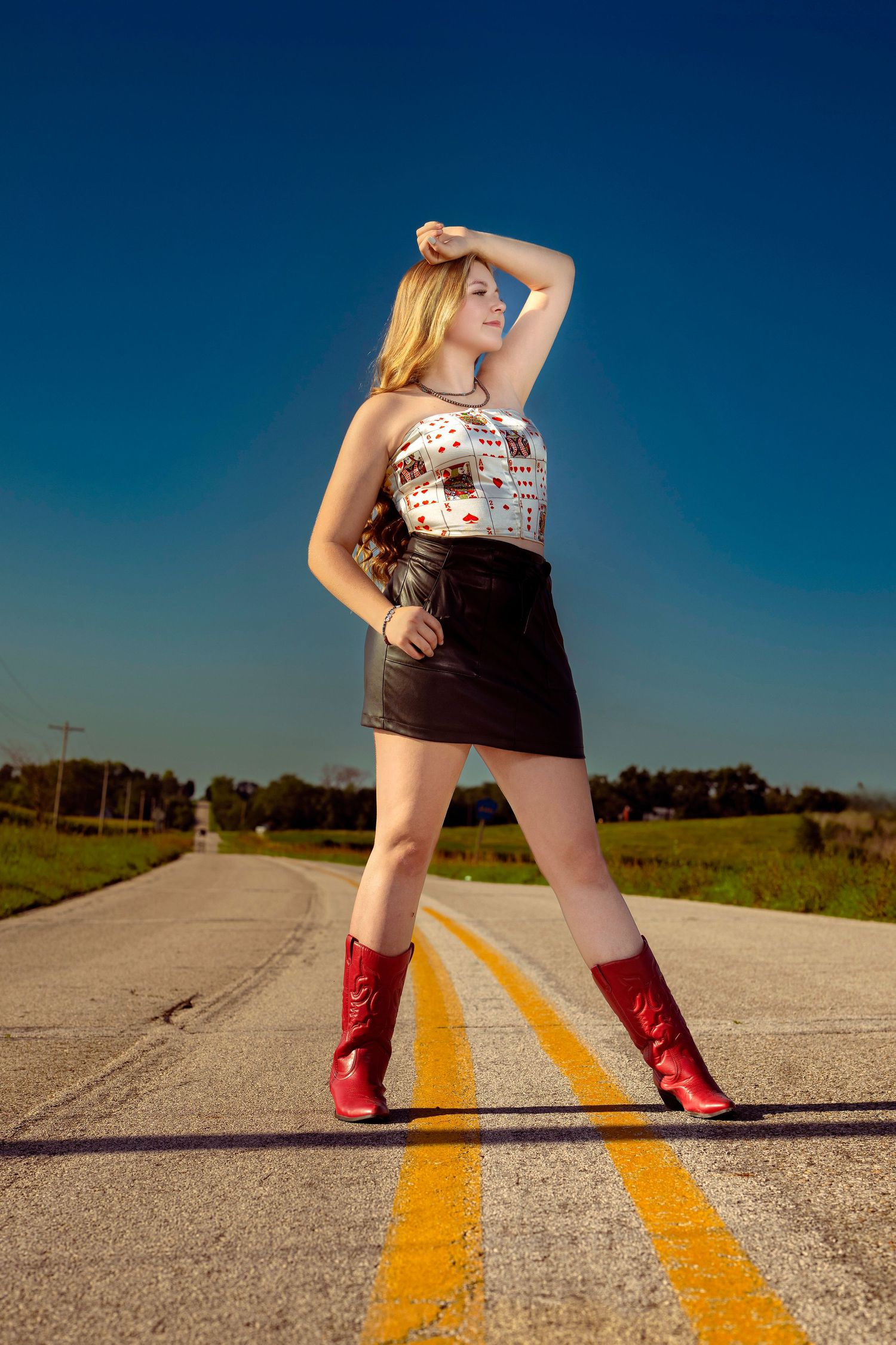 Blonde woman in leather skirt and red cowboy boots posing on rural road.