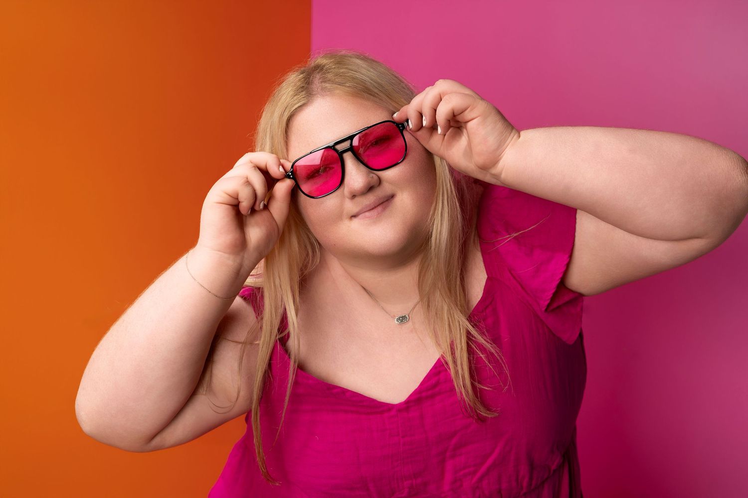 Cheerful blonde woman holding pink sunglasses against bright orange and pink background.