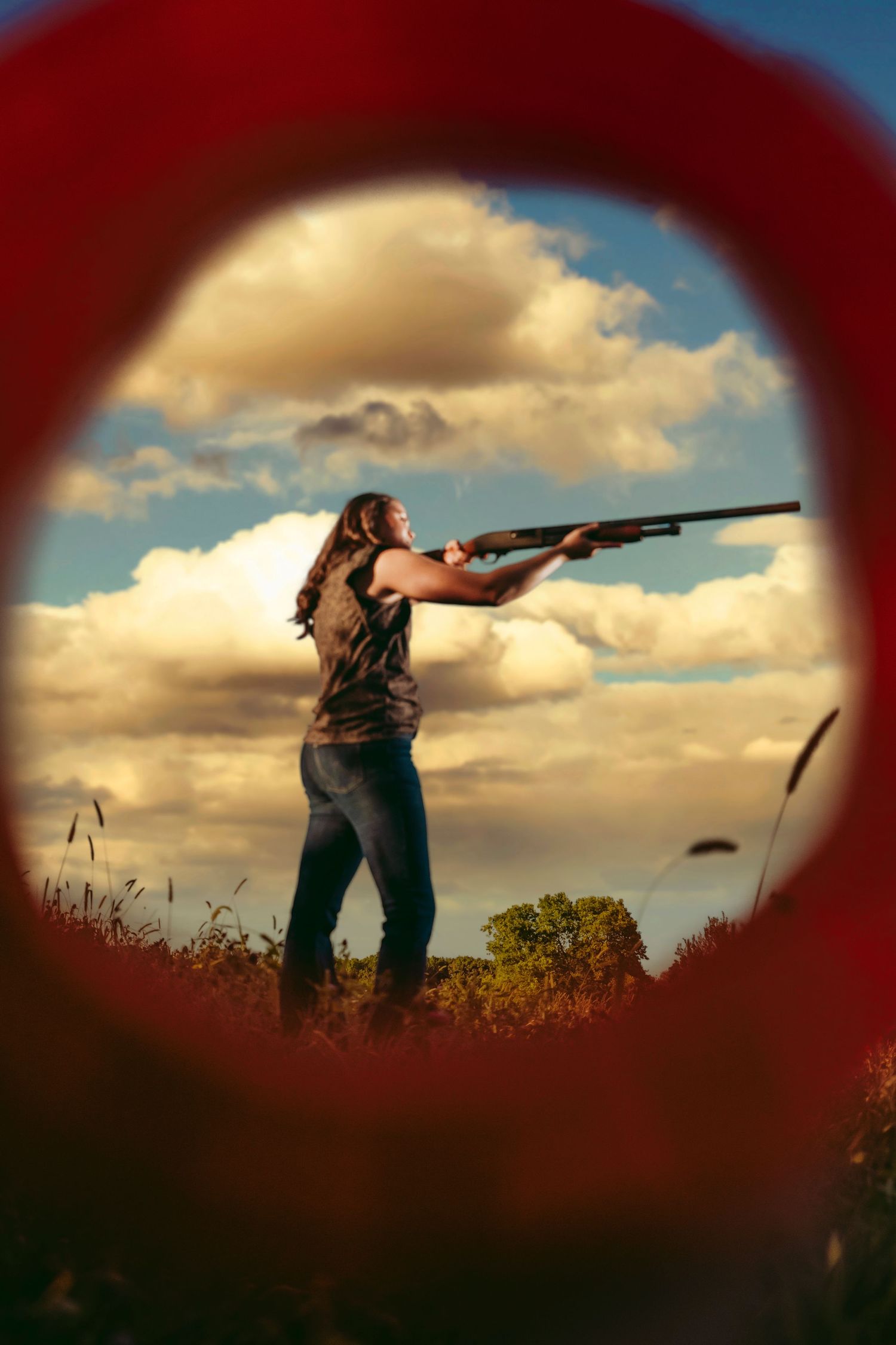 Woman aiming shotgun outdoors framed by red circle with dramatic cloudy sky.