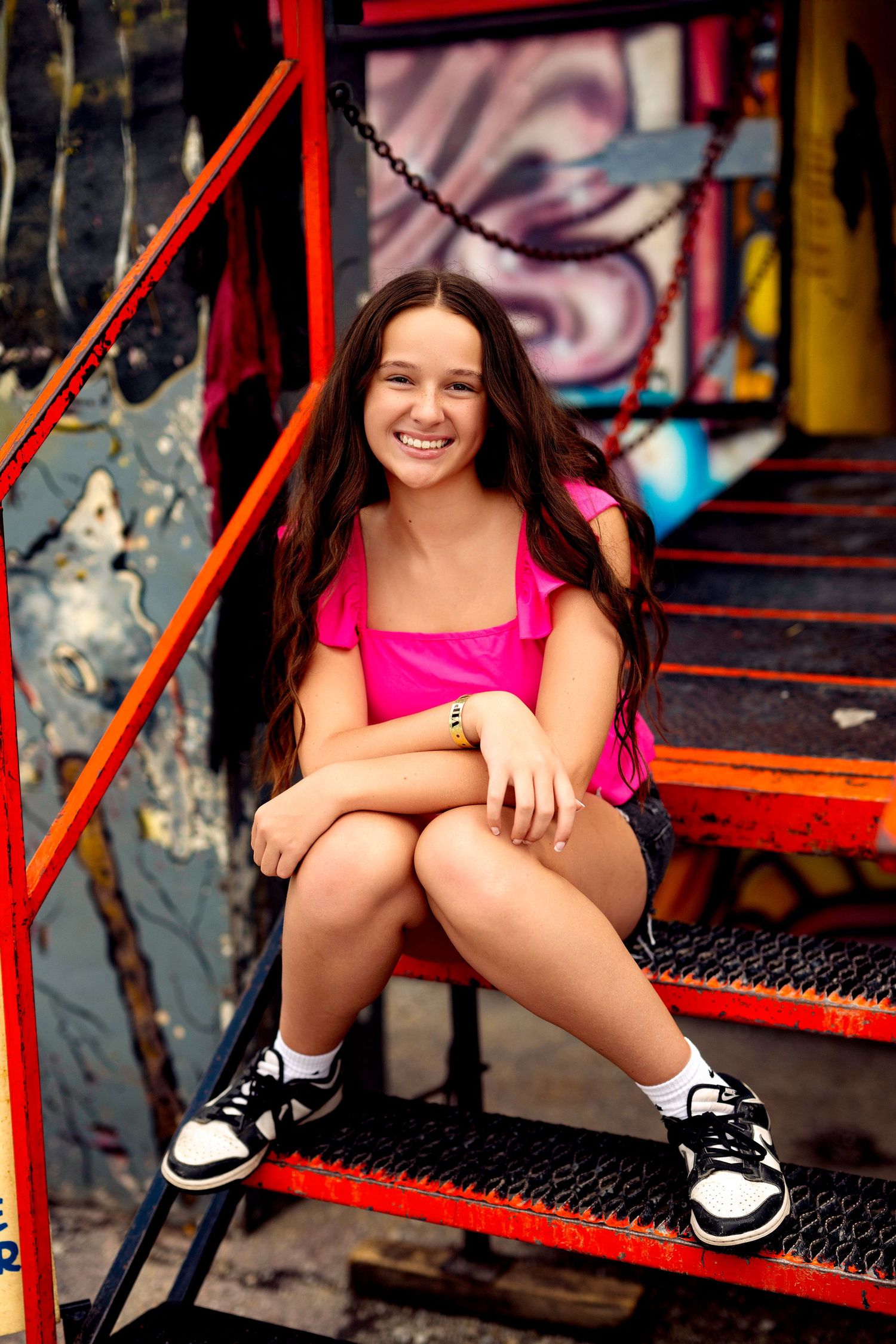 Smiling teen girl in pink top sitting on red metal stairs near graffiti wall. Carnival