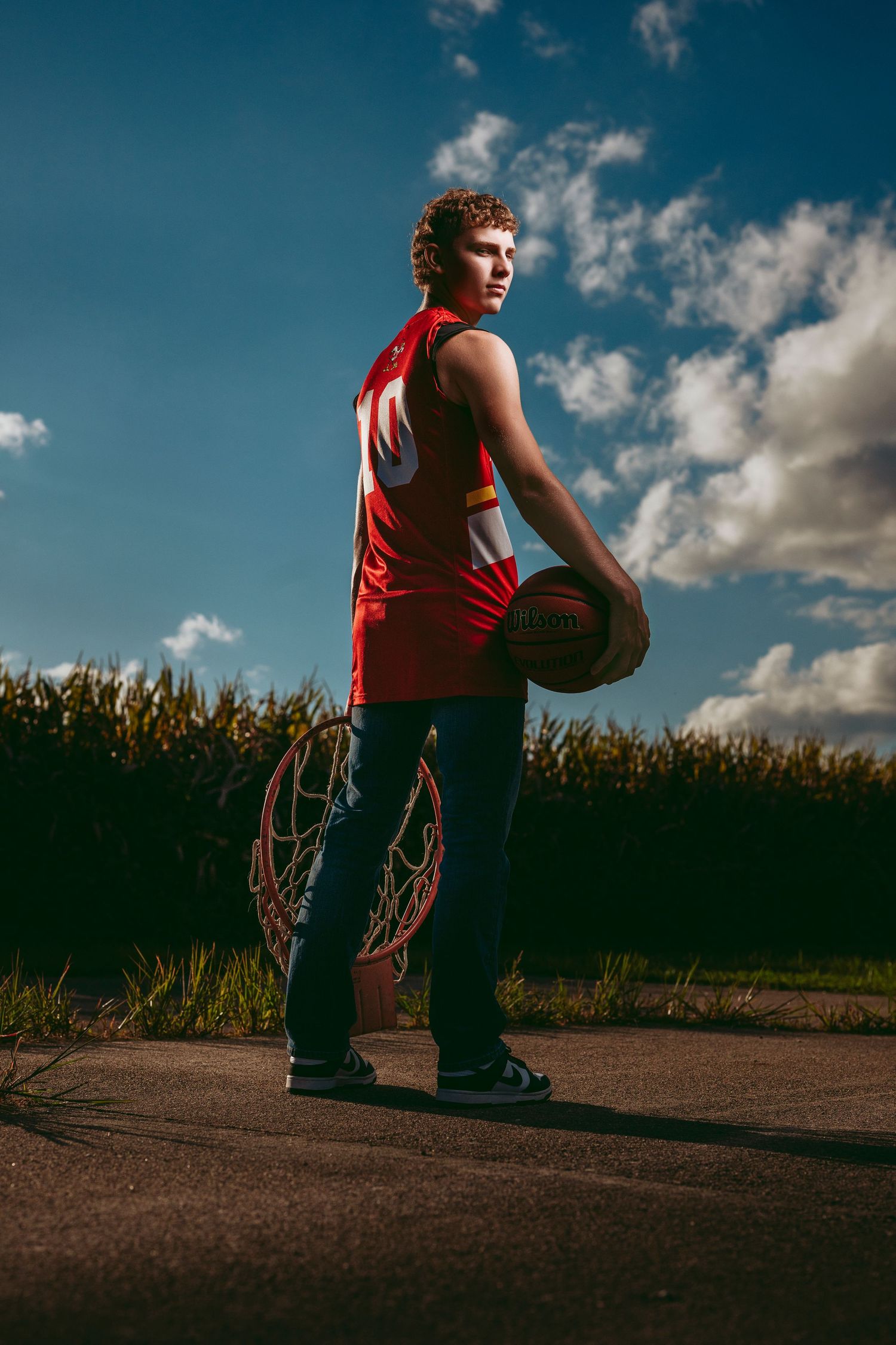 Teen basketball player in red jersey holding ball beside hoop near cornfield under cloudy sky.