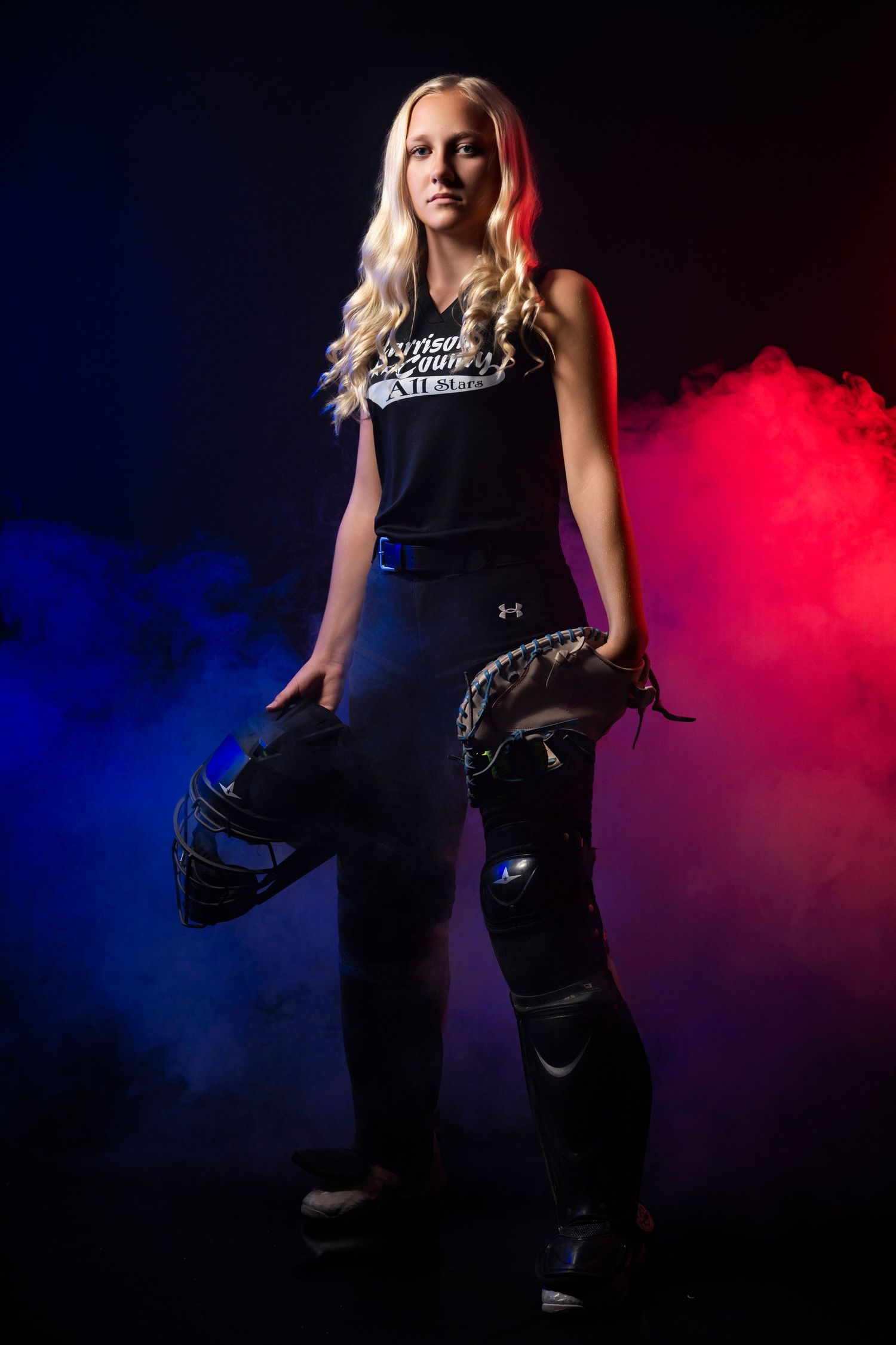 Blonde female softball player in black uniform holding catcher gear against dramatic red and blue smoke background.