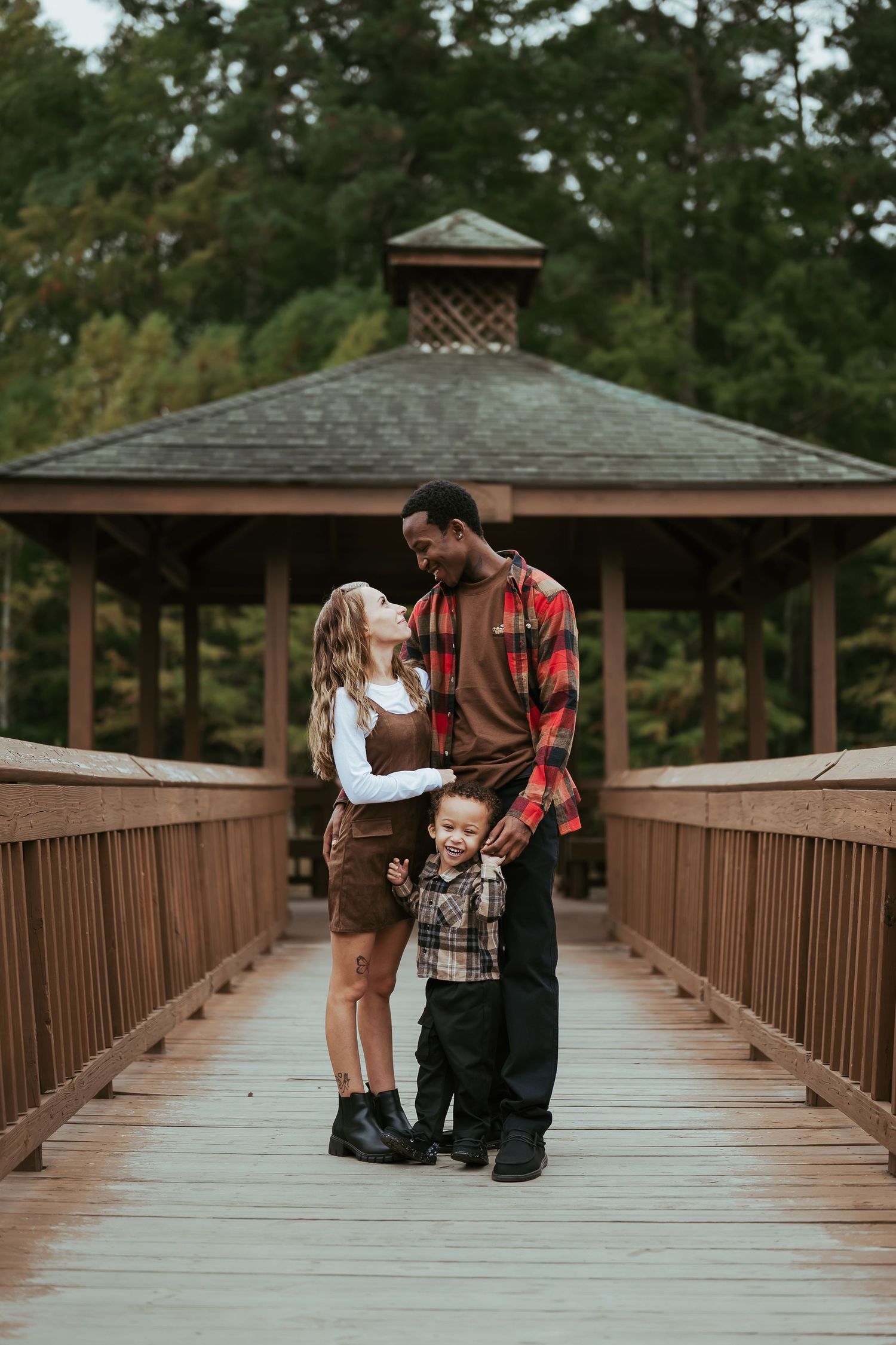 Couple sharing romantic moments on a wooden bridge walkway surrounded by pine trees at sunset.