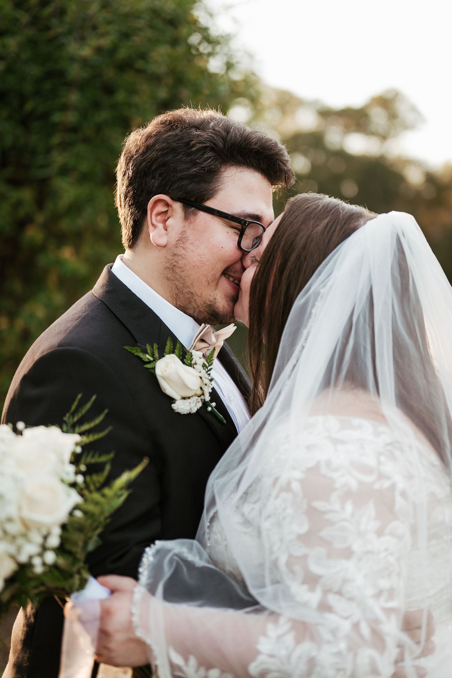 A tender wedding moment captured outdoors with a white boutonniere and lace veil in warm sunlight.