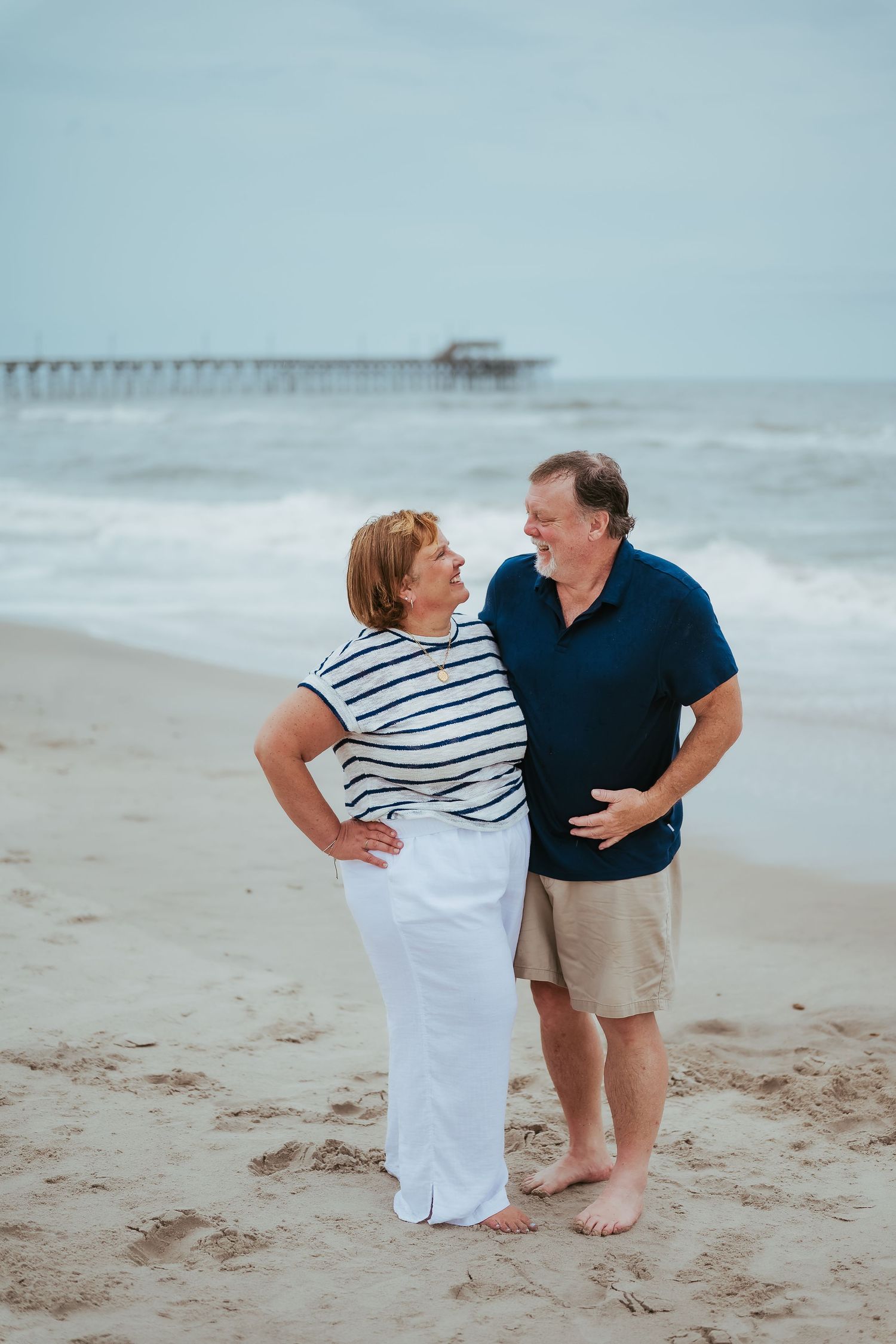 A couple walks together on the beach near a pier, sharing intimate moments in their casual summer outfits.