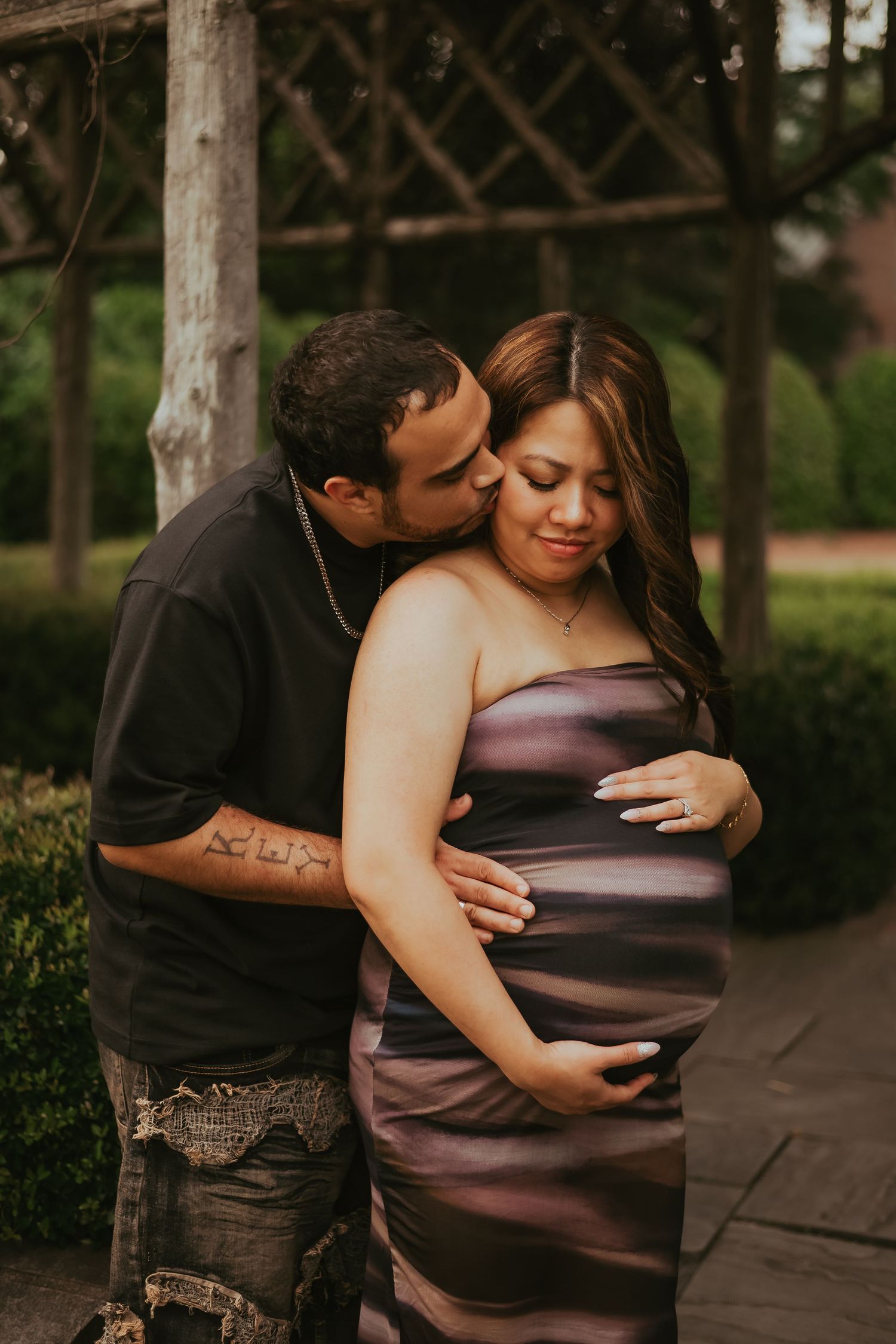 Romantic outdoor maternity photoshoot showing a couple embracing in a park with wooden beams in the background.