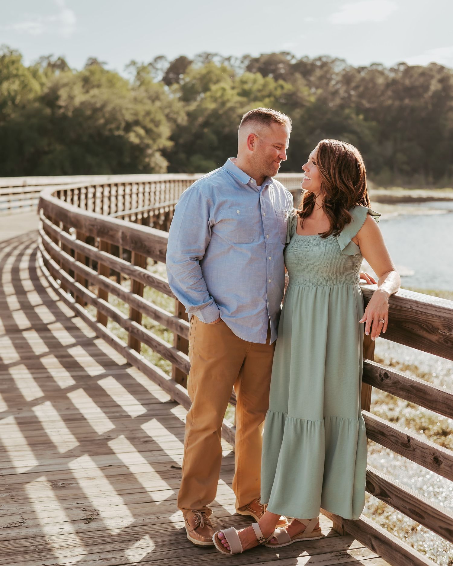 A couple shares a tender moment on a curved wooden boardwalk by a tranquil lake during golden hour.