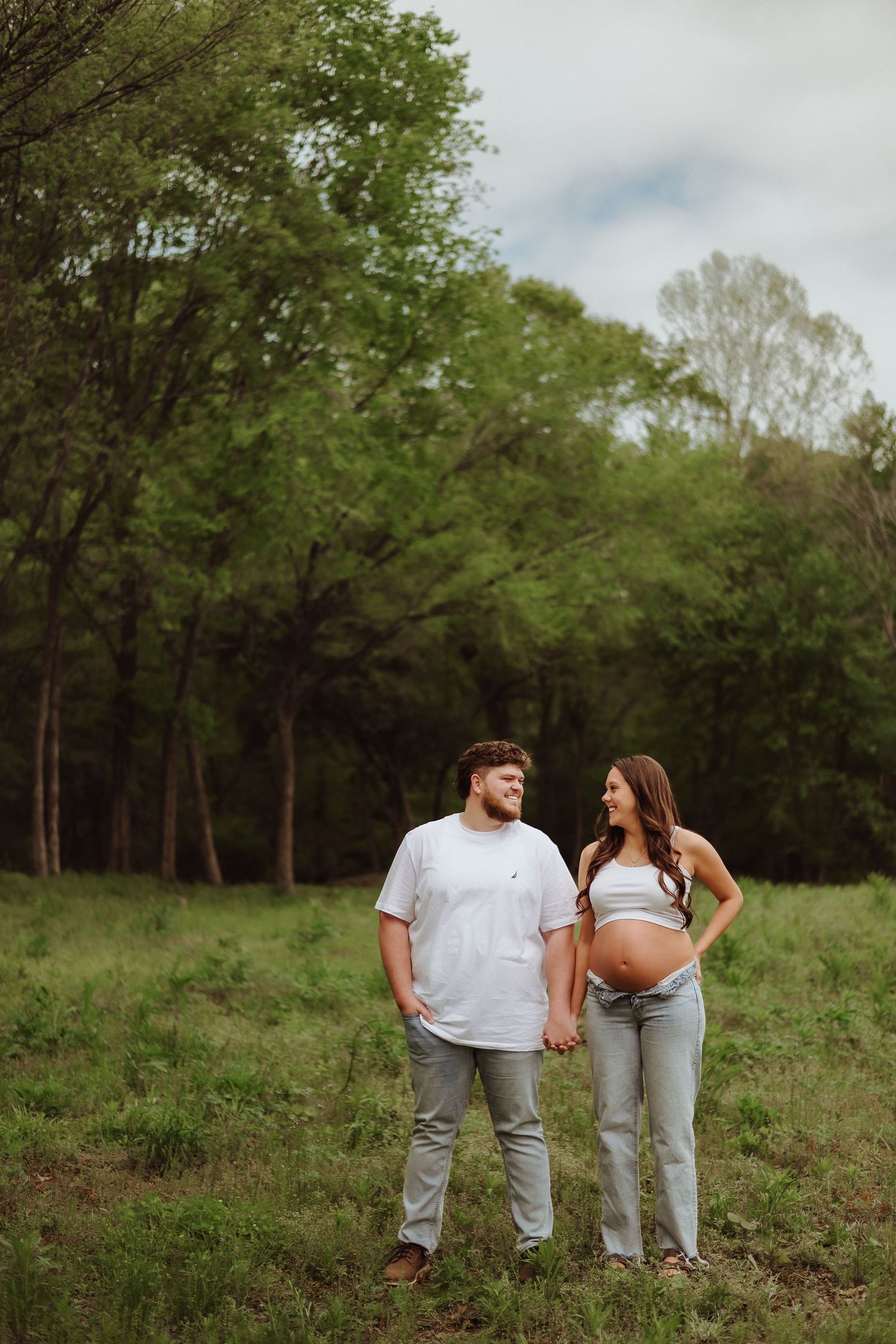 A couple in matching light-colored outfits walk together in a lush green meadow on a cloudy spring day.