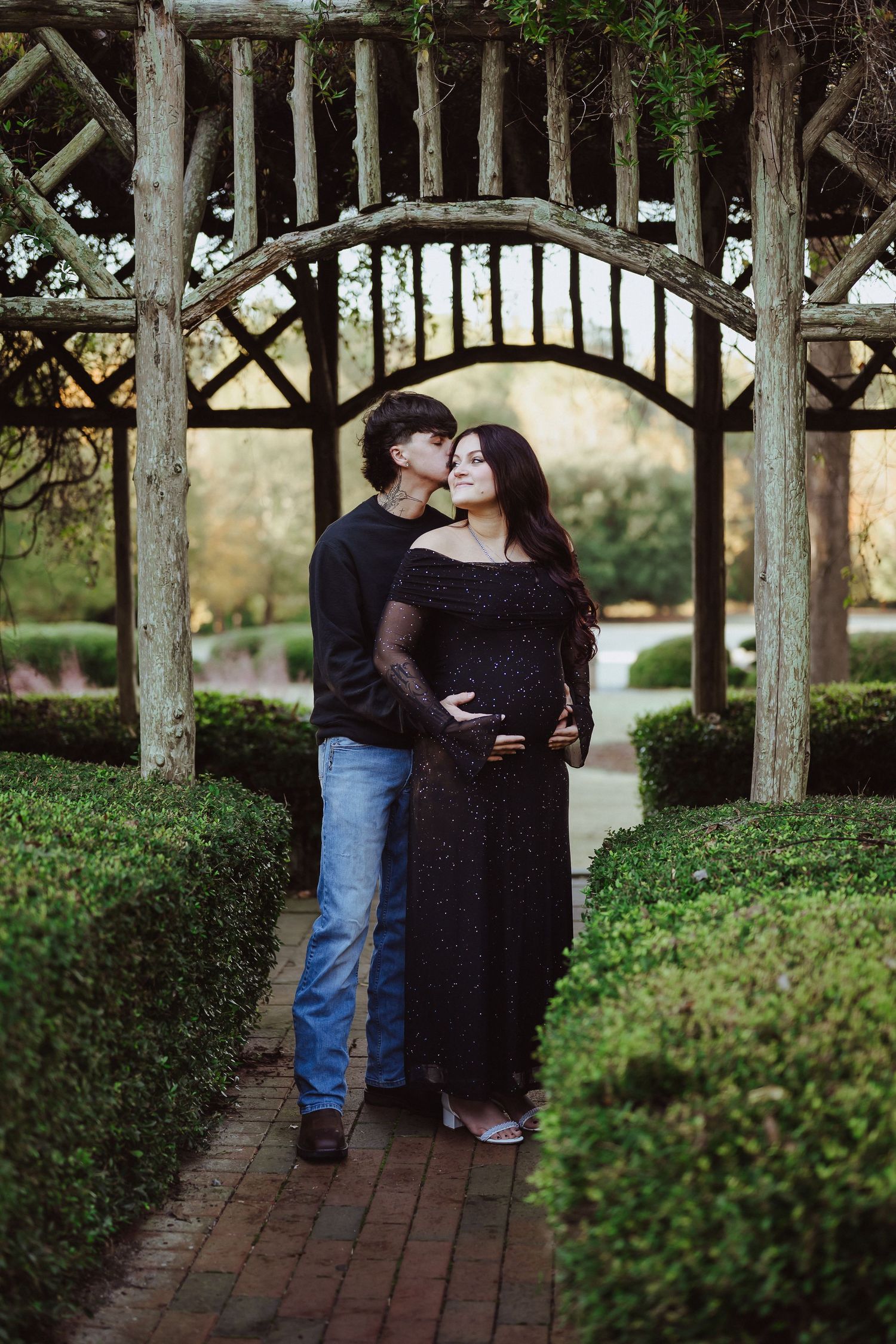 A couple shares a tender moment under a rustic wooden pergola in a garden setting with brick pathways.