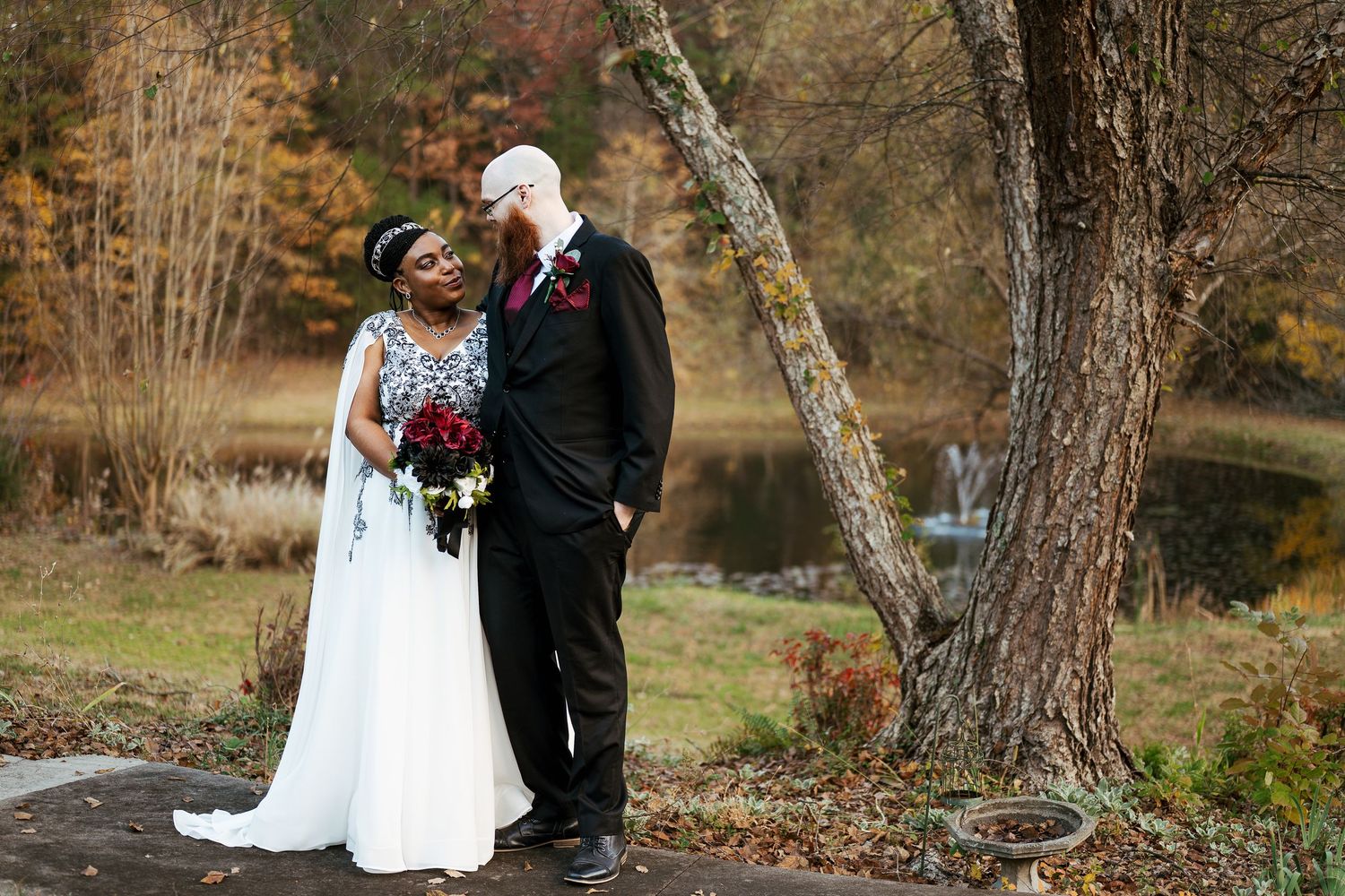 A couple in wedding attire stands together in a scenic autumn setting with trees and fallen leaves.