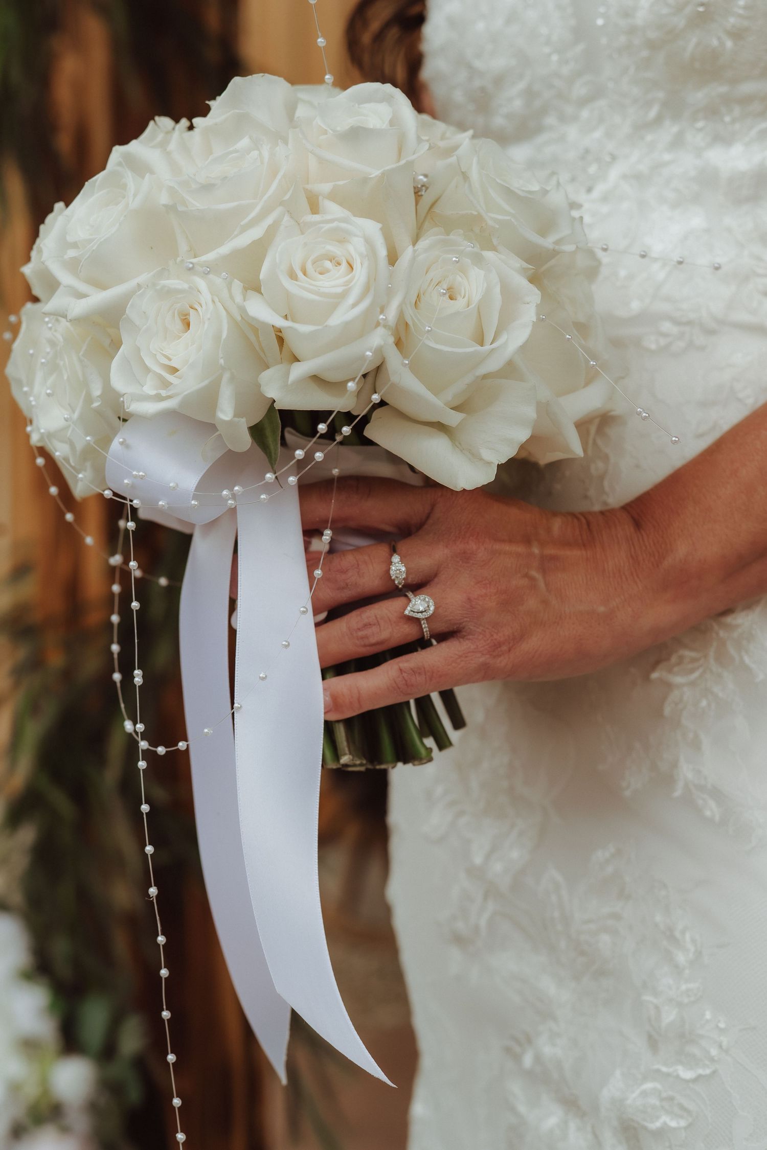 Close-up detail of white rose bridal bouquet with white ribbon and pearl strands held by hands showing engagement ring.