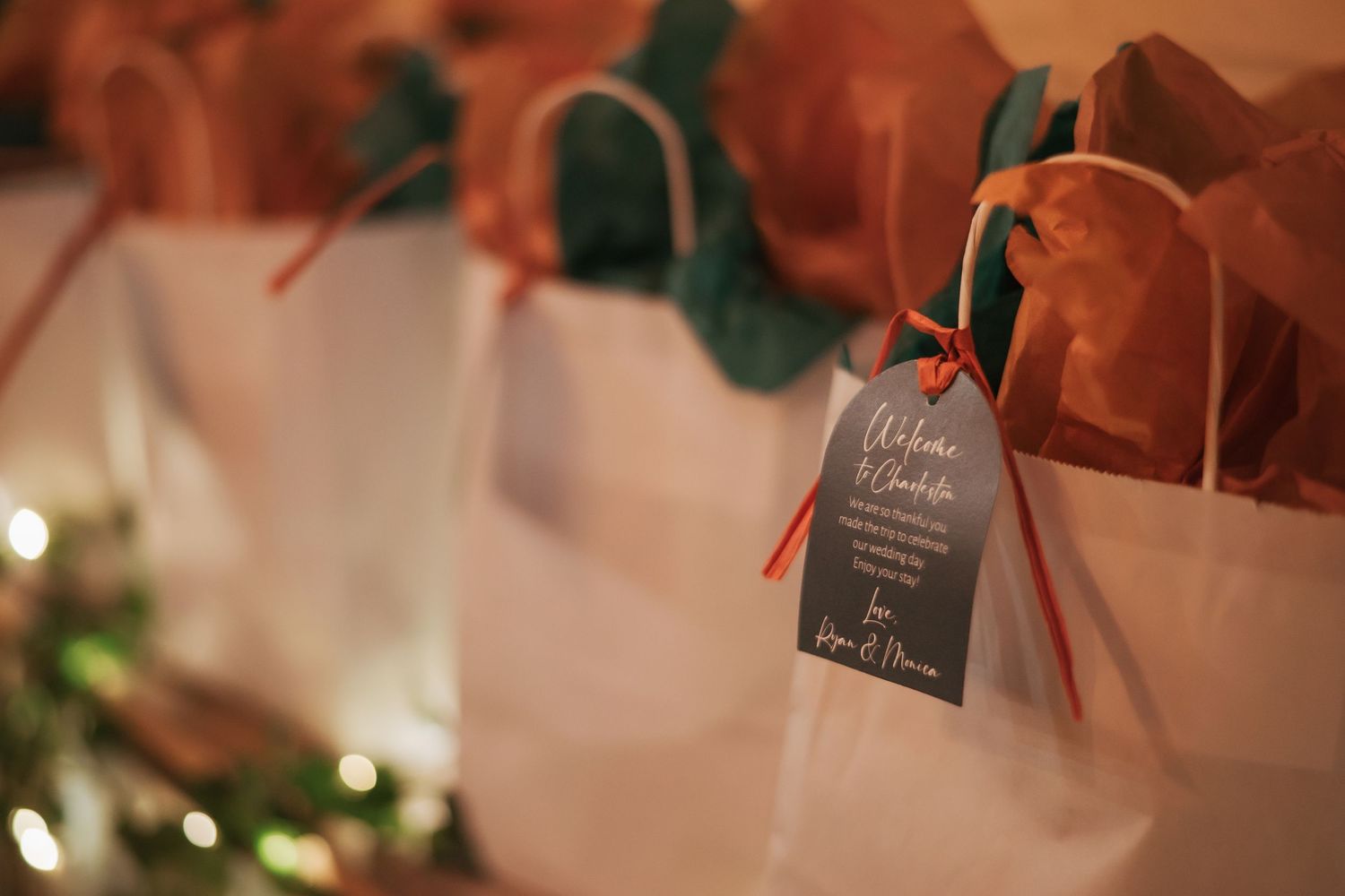 White gift bags with orange tissue paper and small tags arranged in a row with soft lighting in the background.