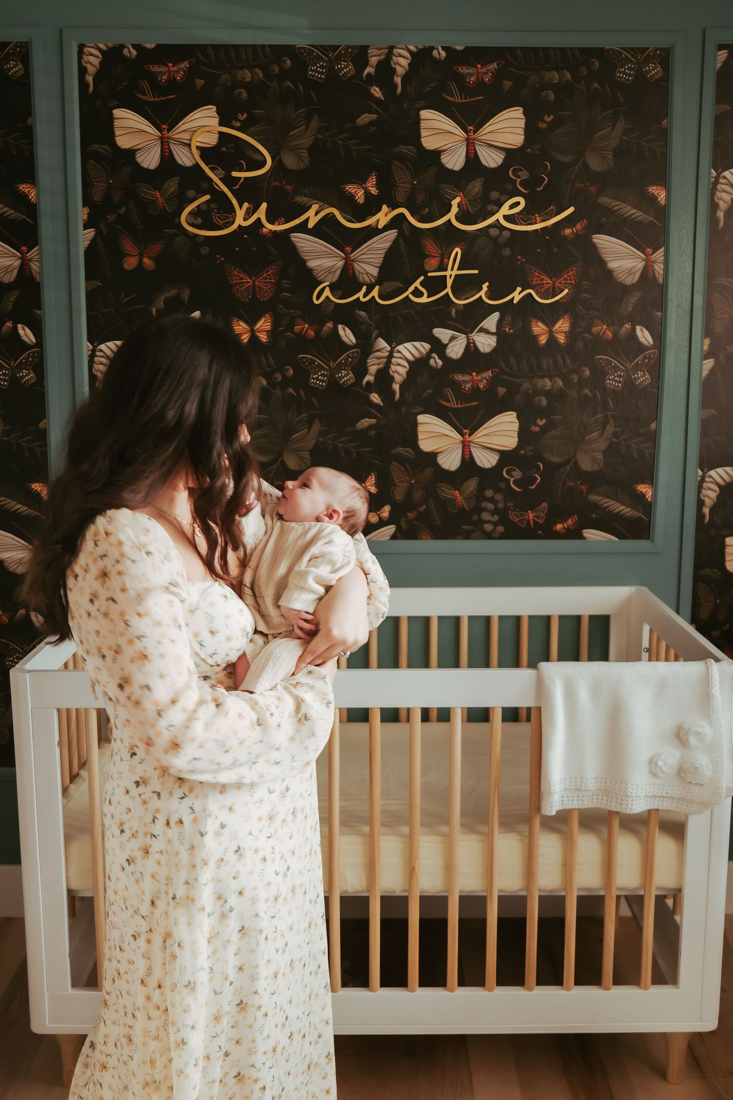 Parent in floral robe placing newborn baby into wooden white crib in nursery with butterfly wall decor.