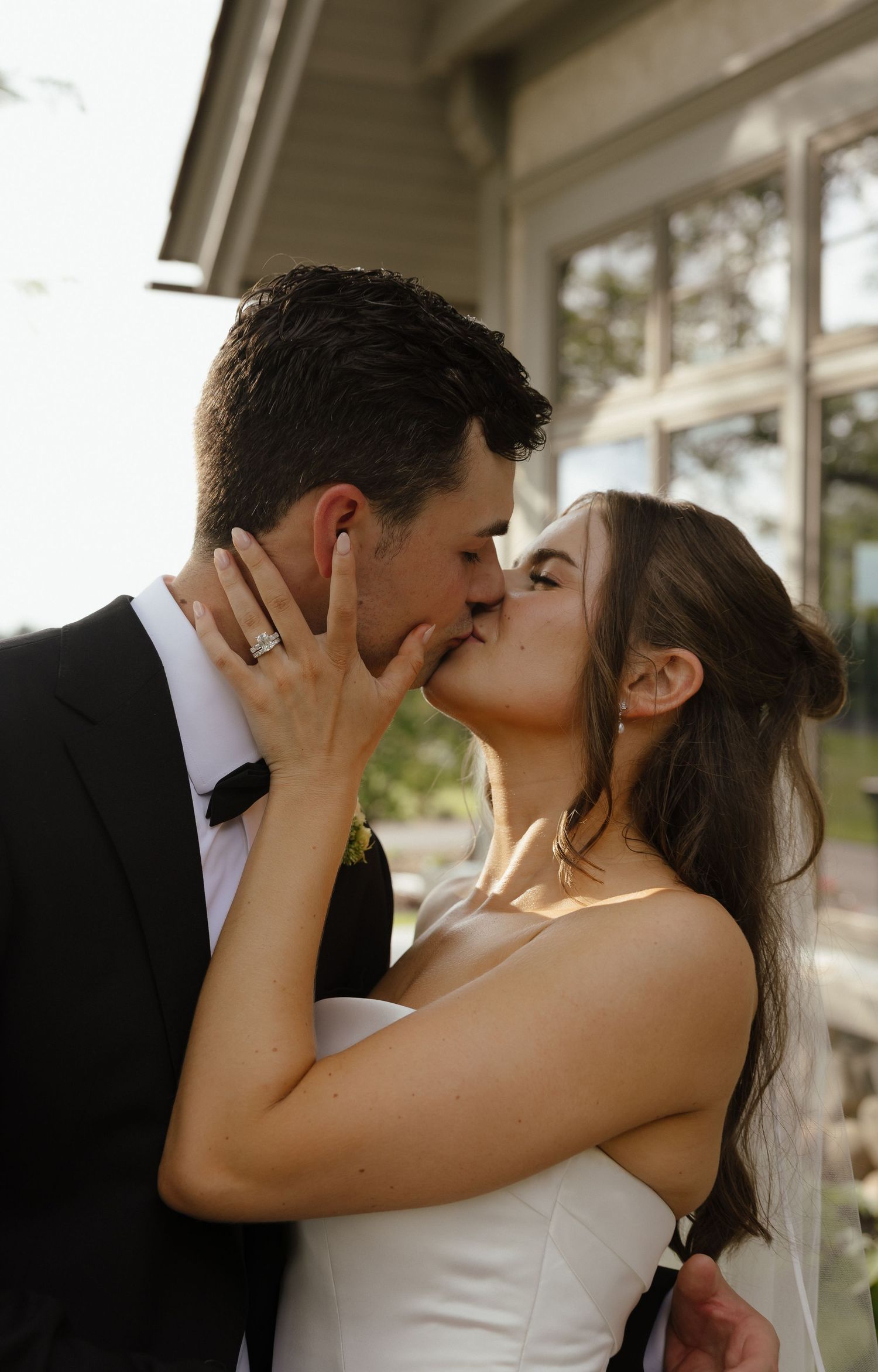 Bride and groom sharing a passionate kiss outdoors, bride wearing diamond ring and white strapless wedding gown.