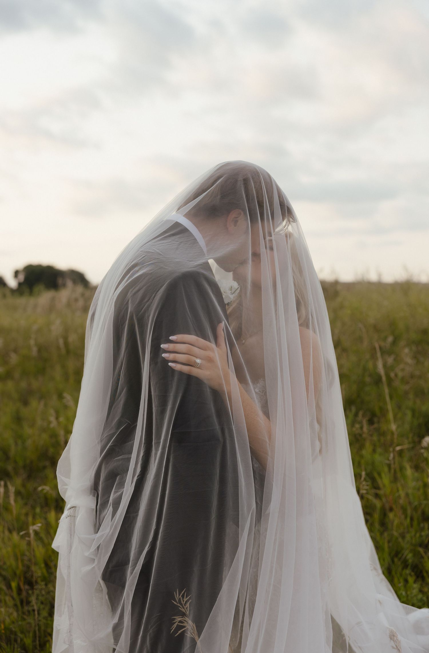 A bride and groom share a romantic kiss under a flowing white veil in a lush green field on their wedding day.