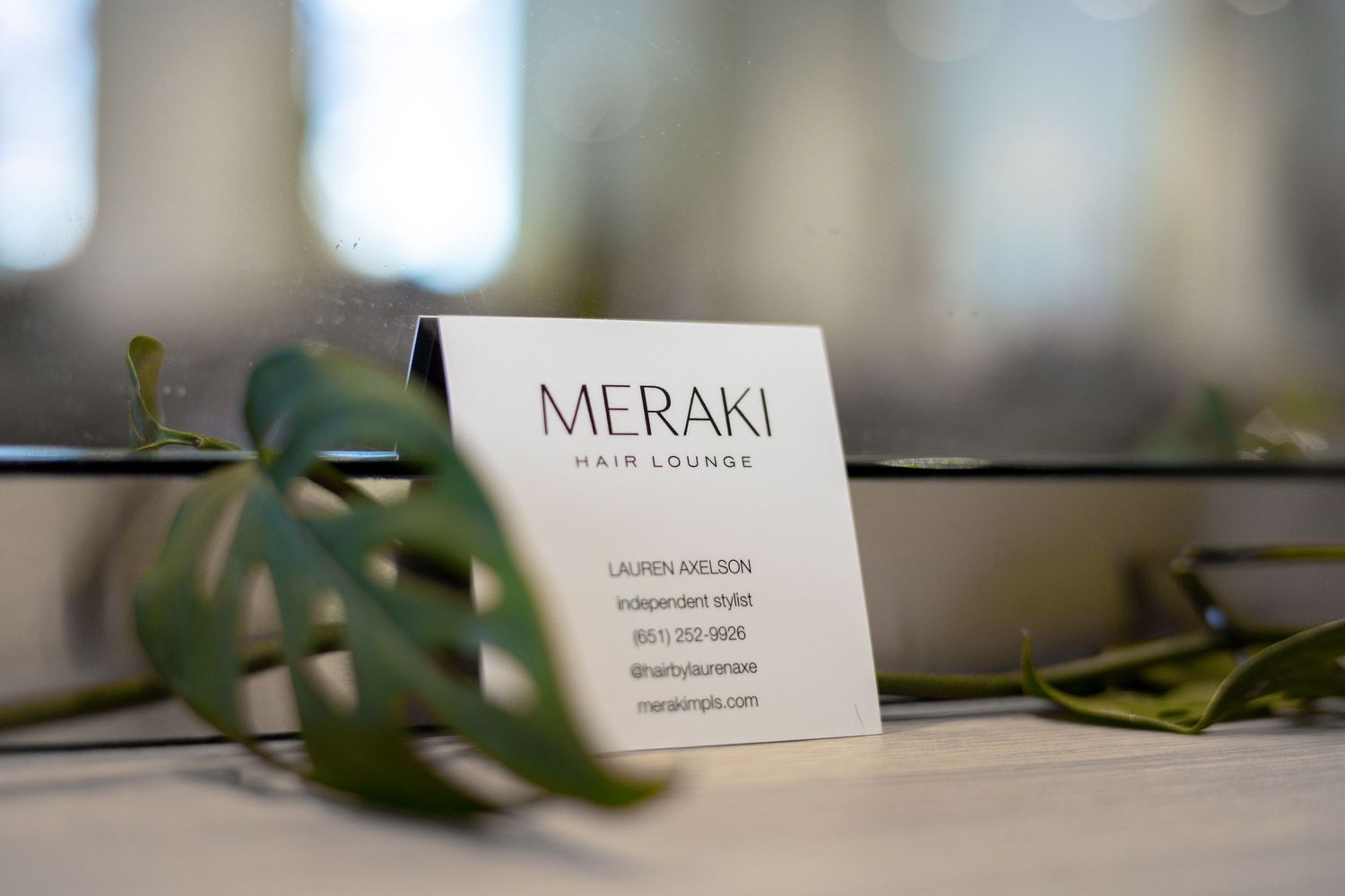 Meraki Hair Lounge business card displayed with tropical green leaves in a modern salon.