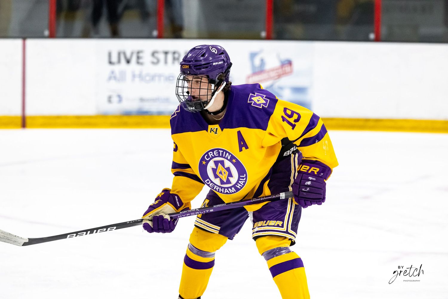 Hockey player wearing purple and gold uniform with number 19 during ice hockey game.