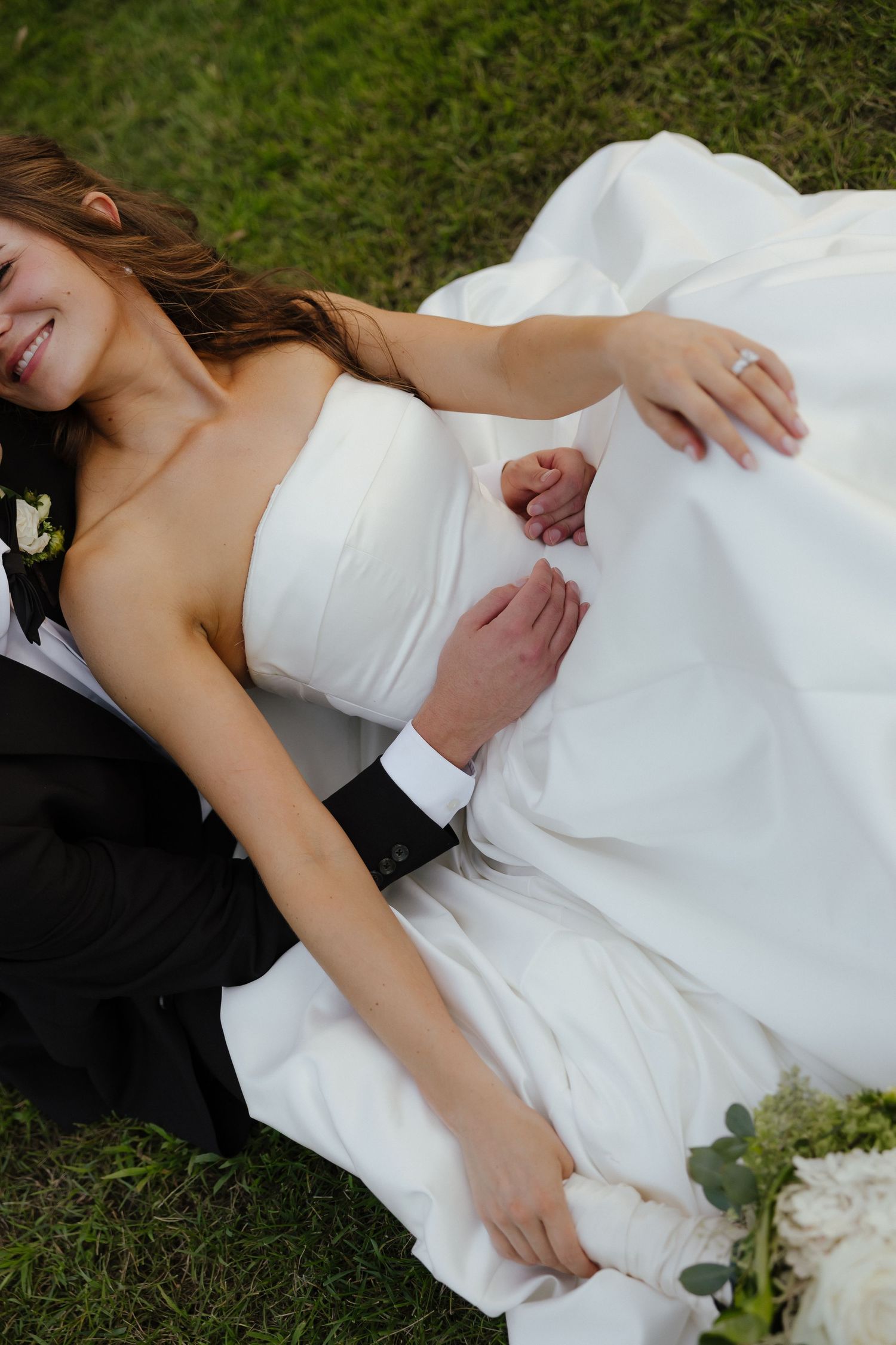 Bride and groom lying on grass holding hands, bride in strapless white gown with white floral bouquet nearby.