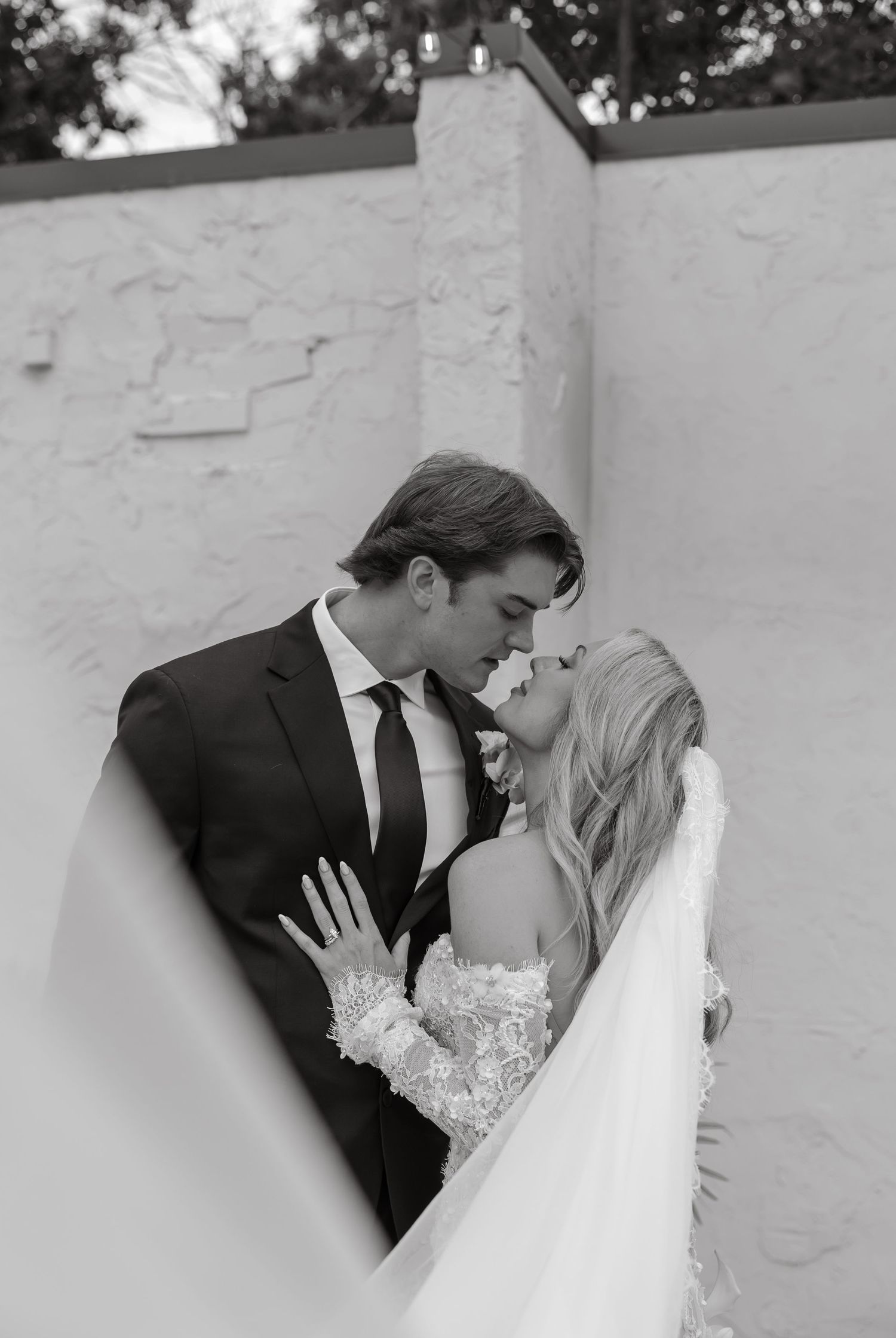 Black and white photo of bride and groom sharing a romantic kiss against a white textured wall.