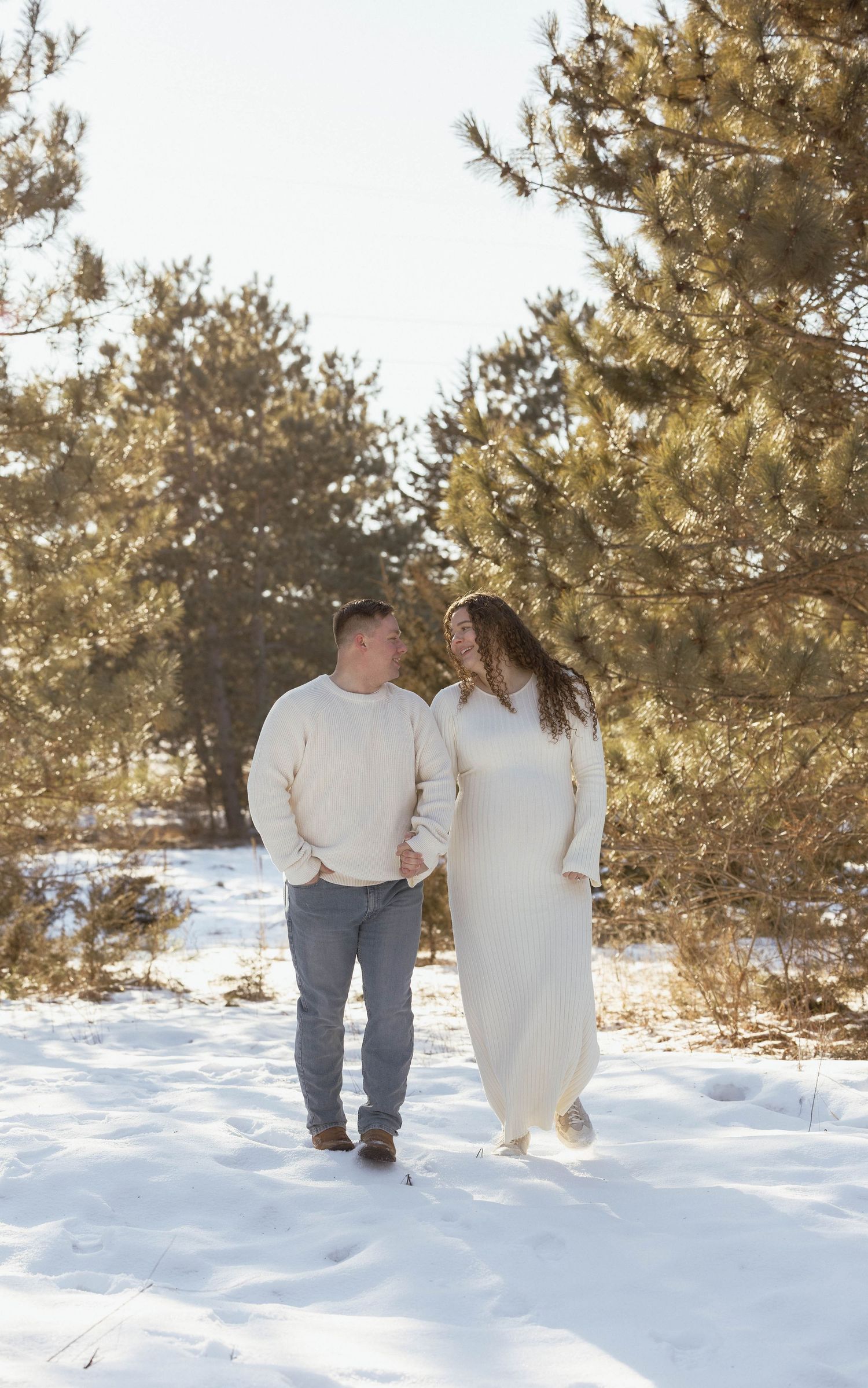 Expecting couple holding hands and walking through a snowy winter forest in matching cream outfits.