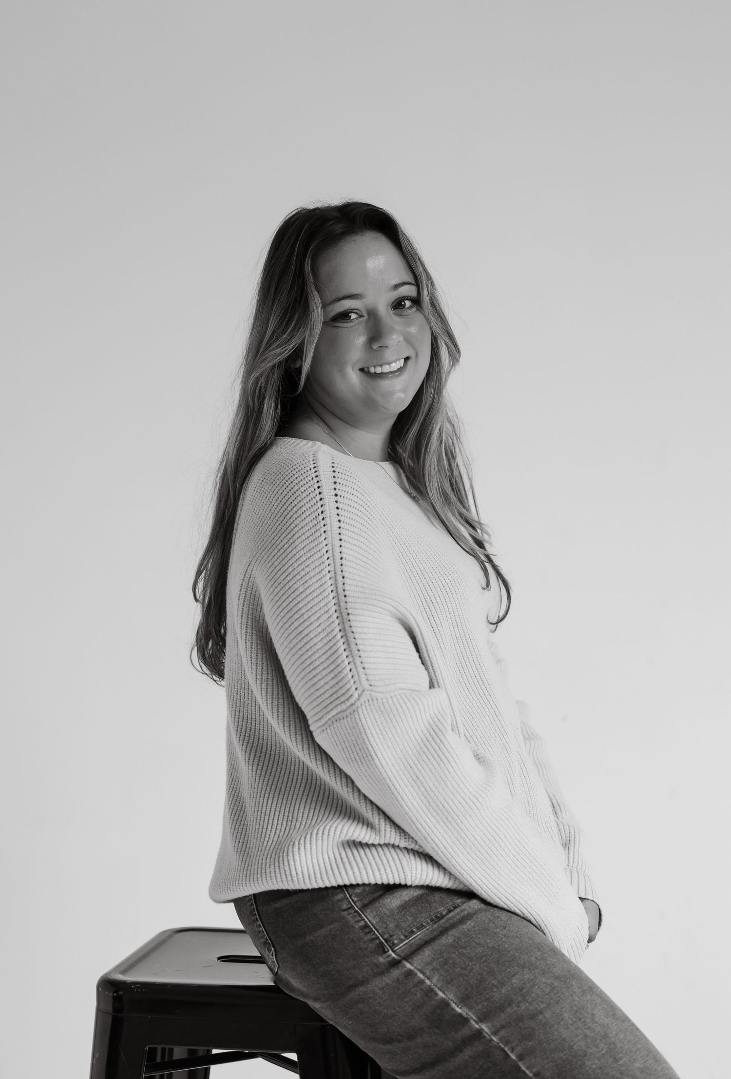 Black and white photo of smiling woman in knit sweater sitting on stool.