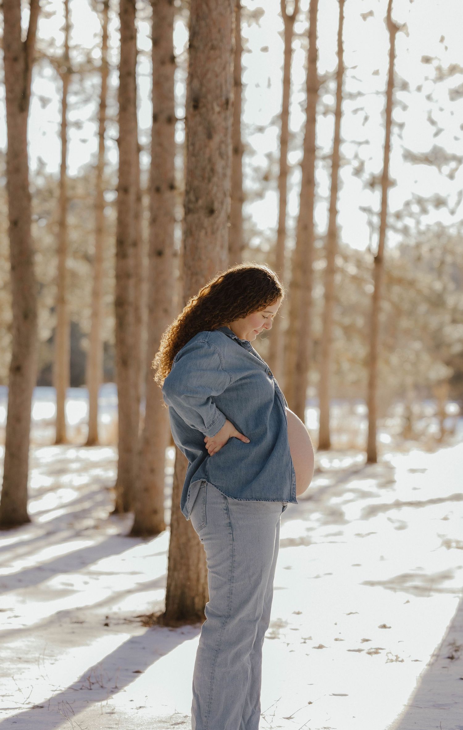Side profile of a pregnant woman in a denim shirt standing among tall trees in a snowy winter forest.