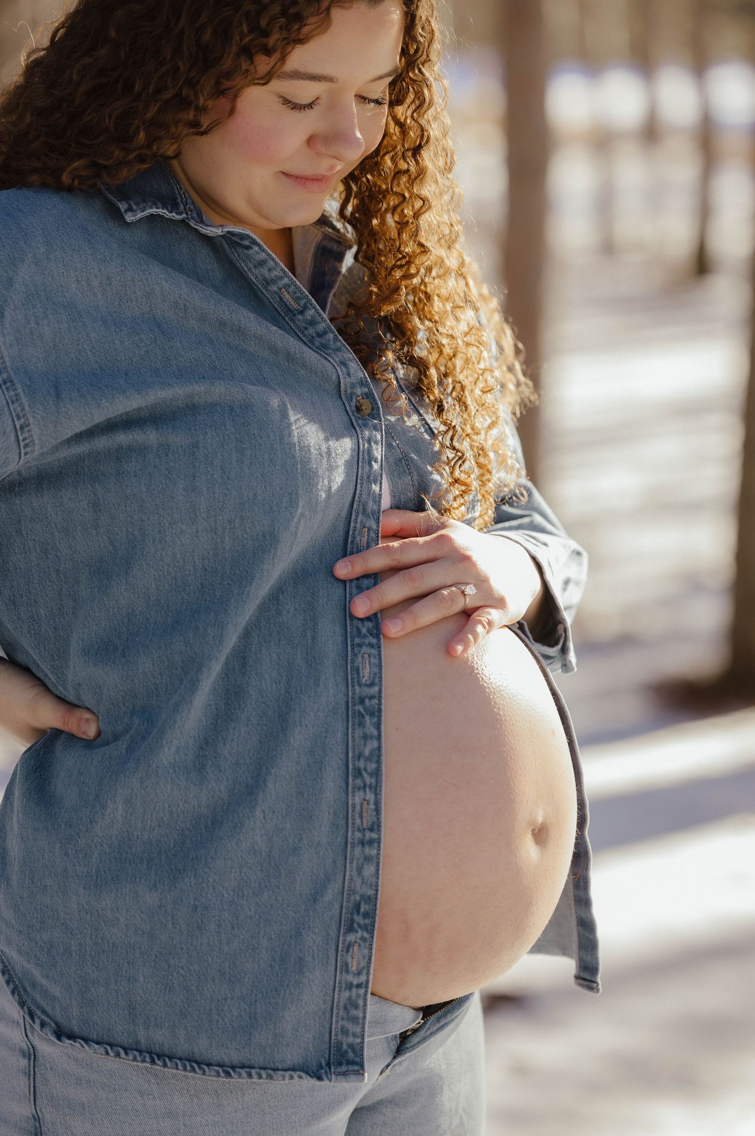 Pregnant woman in denim shirt cradling her baby bump outdoors in sunlight.
