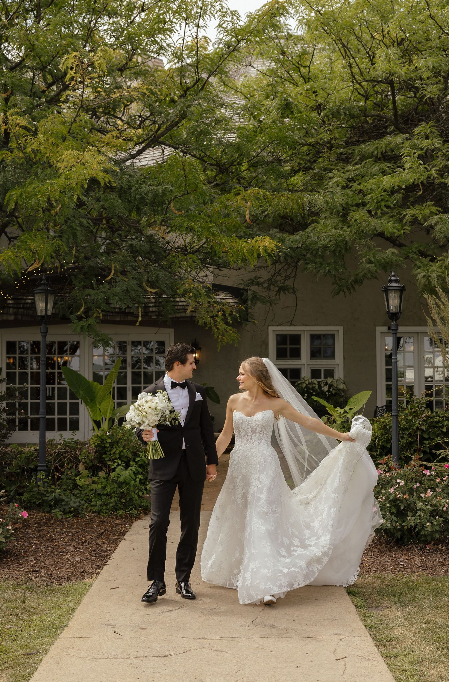 Bride and groom walk hand in hand along garden path surrounded by lush green trees.