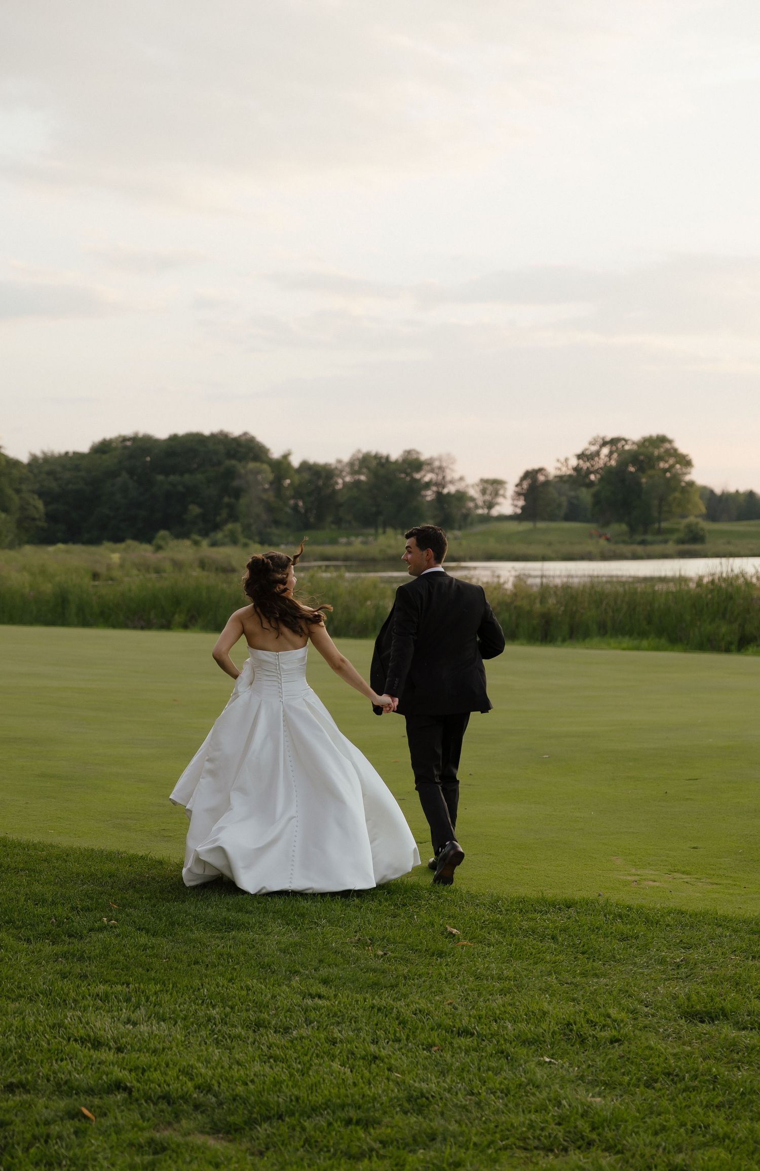 Bride and groom walking hand in hand across a lush green golf course toward a serene lake at dusk.