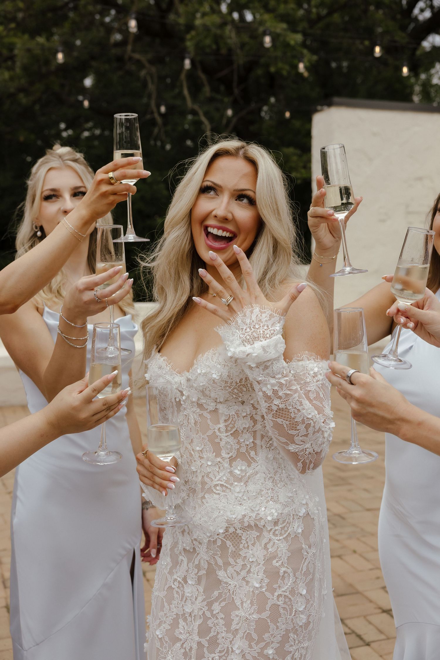 Excited bride in lace gown celebrating with bridesmaids raising champagne glasses outdoors.