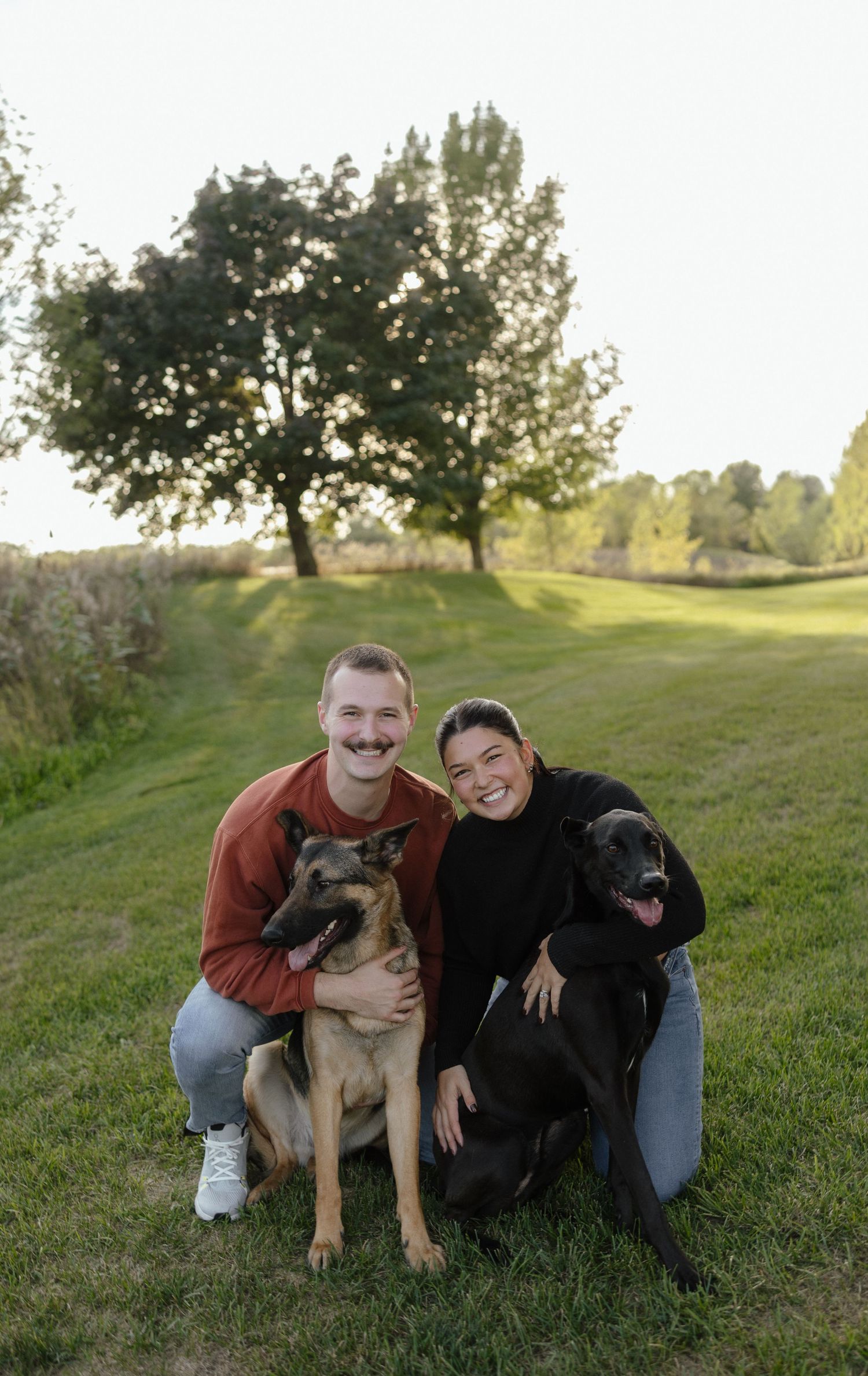 Smiling couple kneels on green grass with their German Shepherd and black Labrador dogs at sunset.