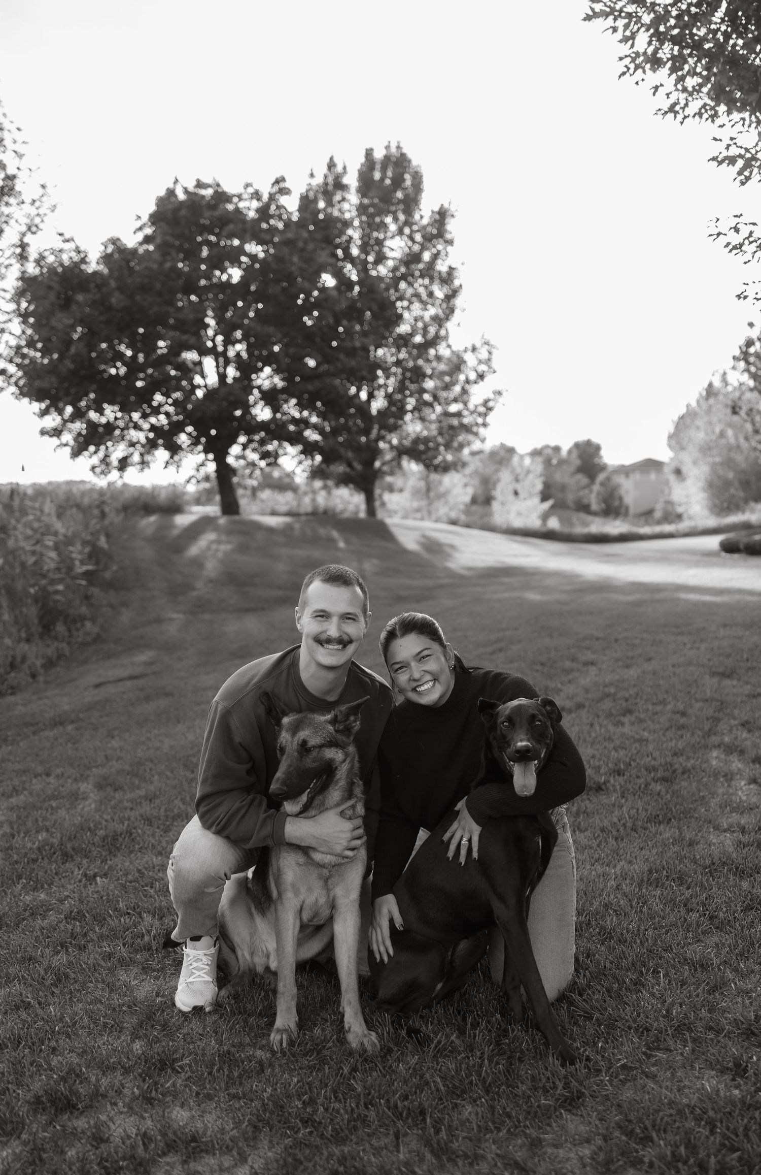 Smiling couple poses outdoors with two dogs on a grassy lawn in black and white photo.