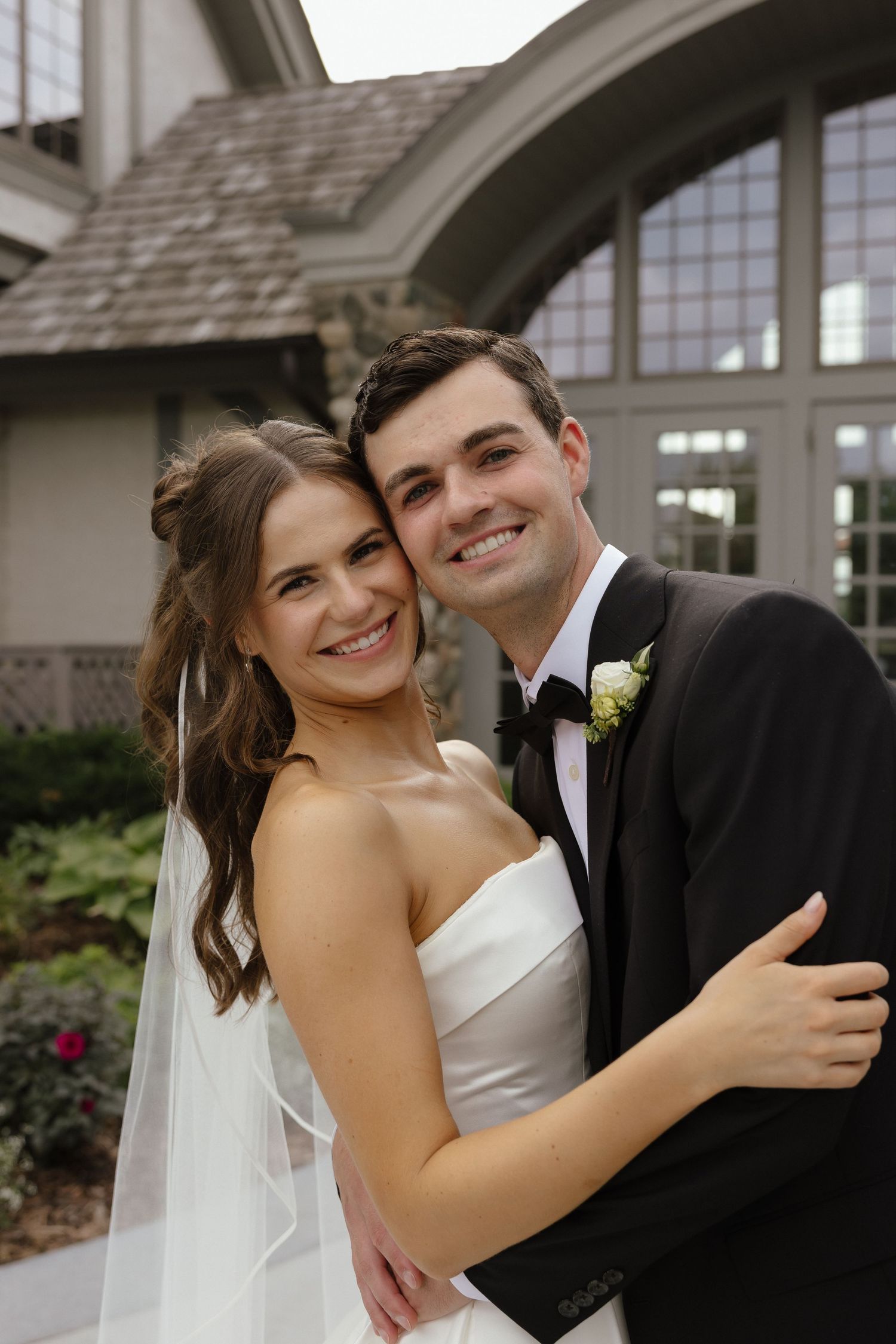 Smiling bride and groom embracing outside stone and wood wedding venue, bride in strapless gown with veil.