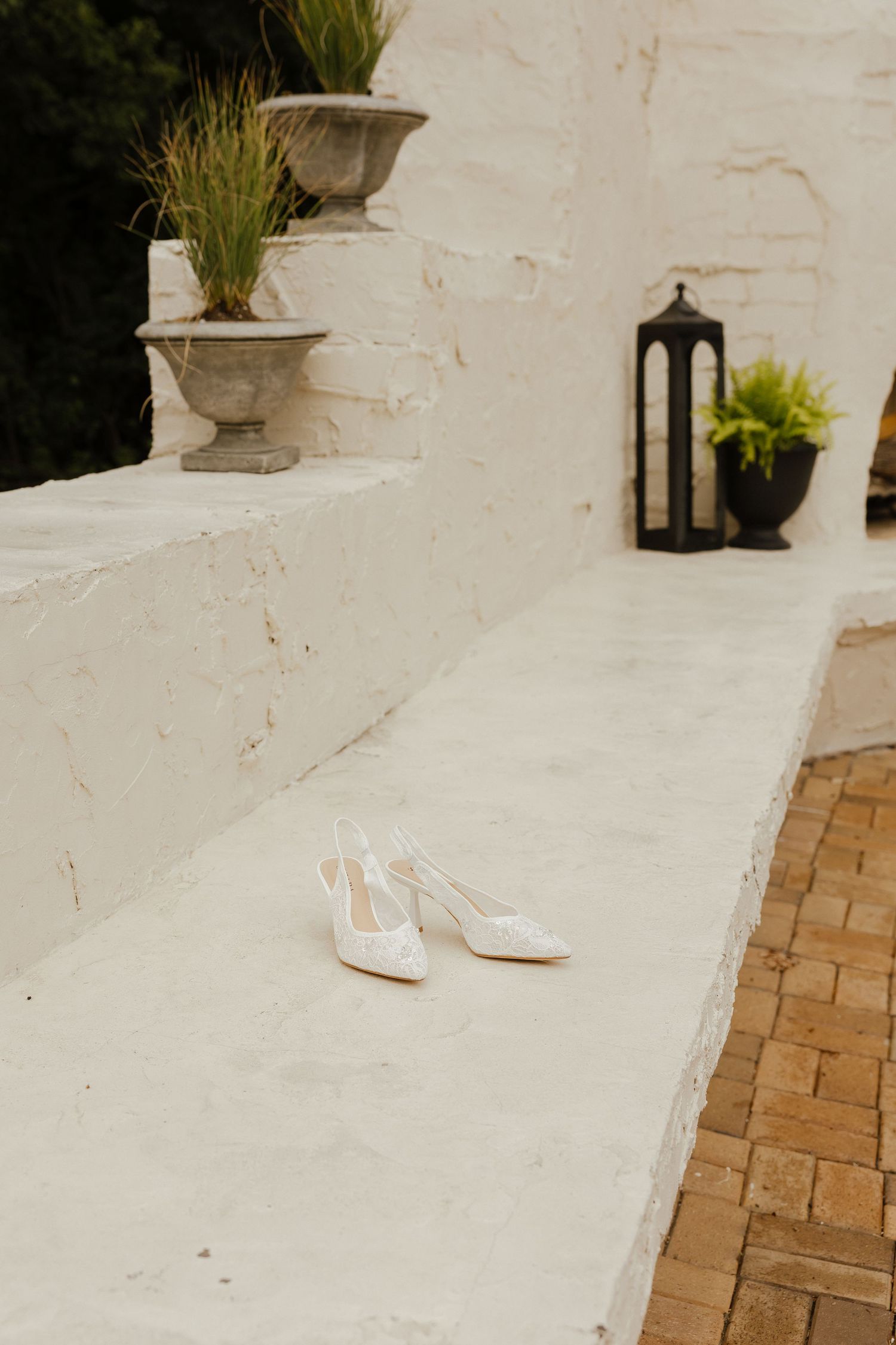 White lace bridal heels placed on a white stone ledge with decorative urns and lantern backdrop.