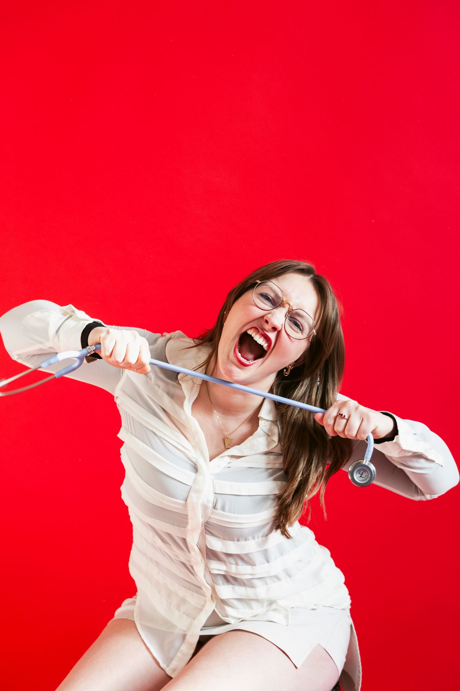 Expressive woman in glasses pulling stethoscope apart while screaming against a vibrant red background.