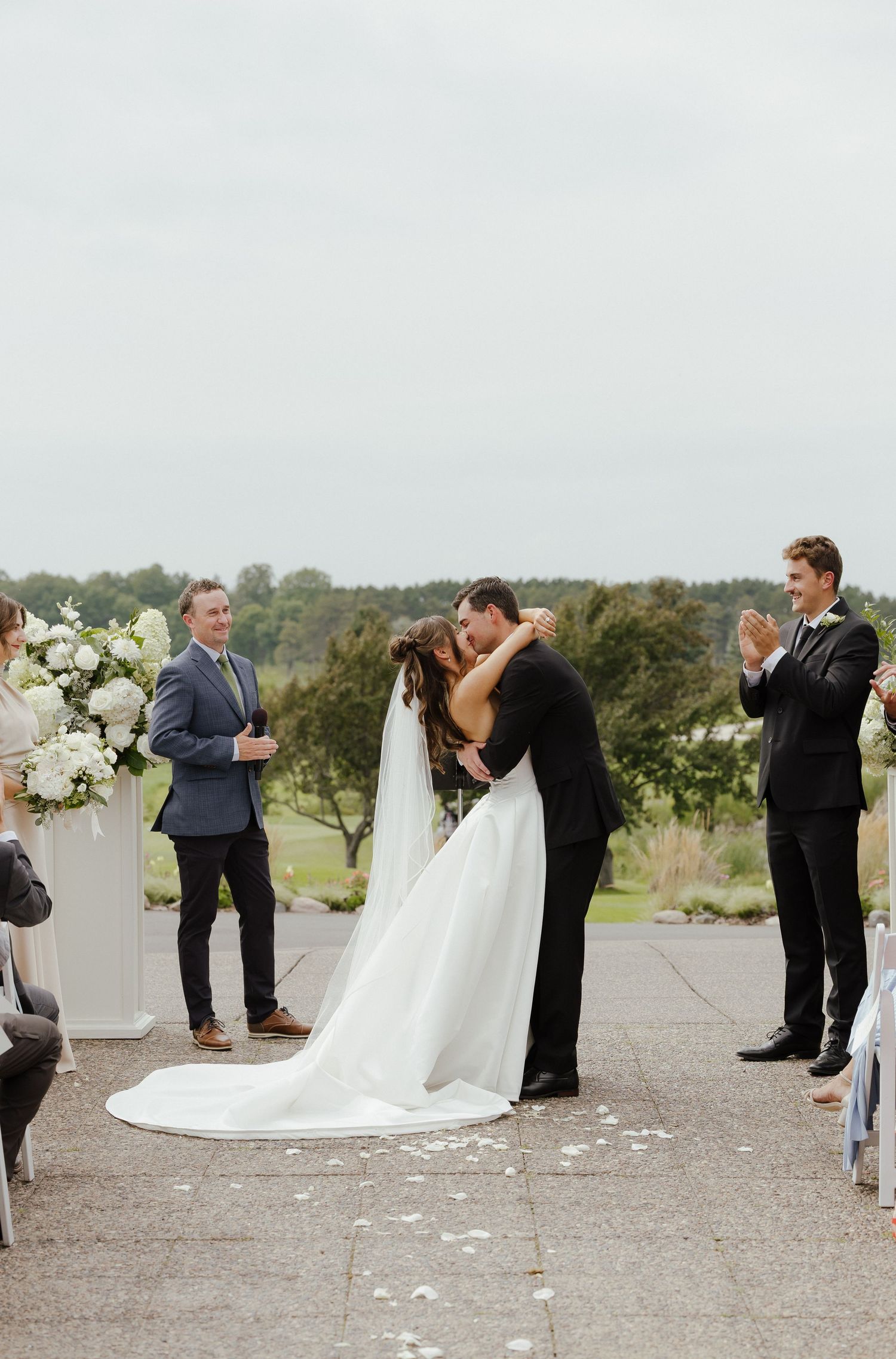 Bride and groom share first kiss at outdoor ceremony altar as wedding party applauds on overcast day.
