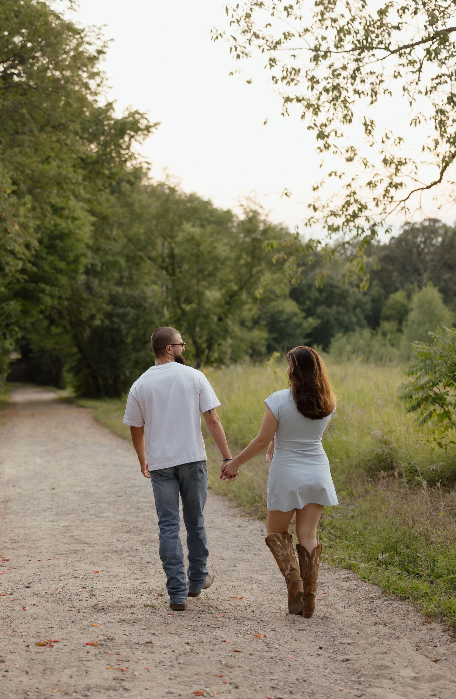 Couple holding hands walking down a rustic dirt path surrounded by lush green trees, woman wearing white dress and cowboy boots.