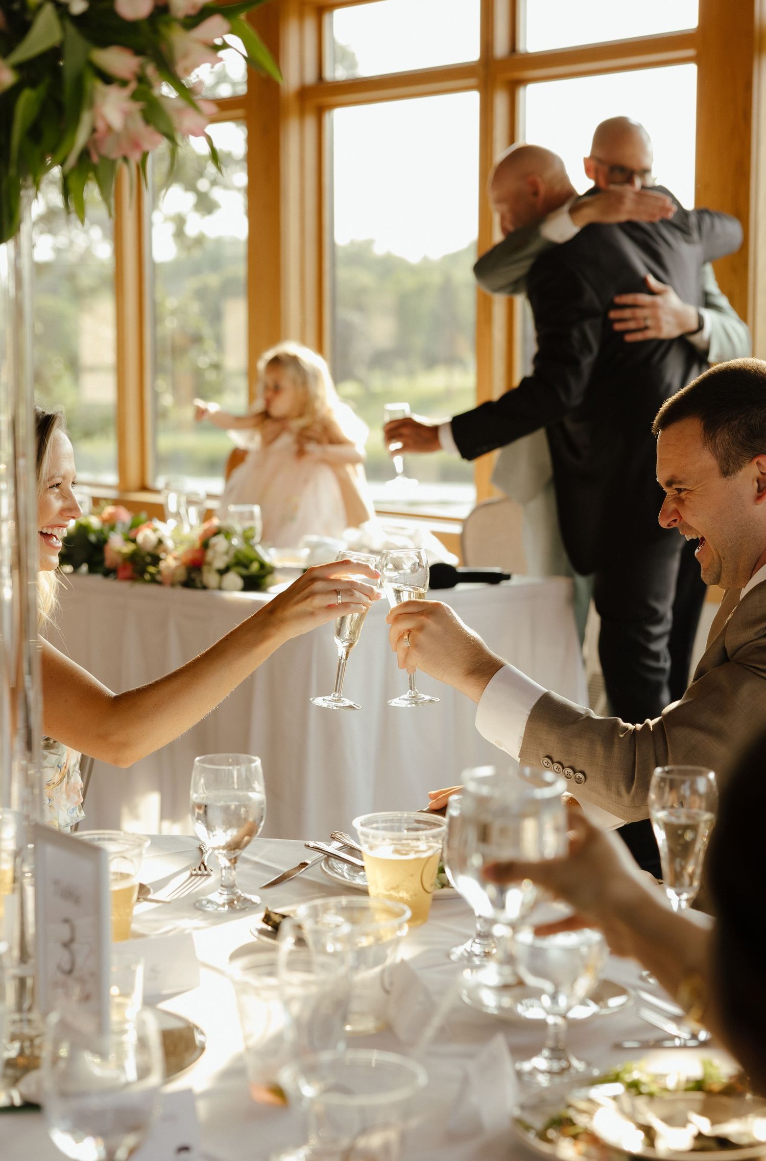 Bride and groom toast champagne at wedding reception table with guests celebrating behind them.