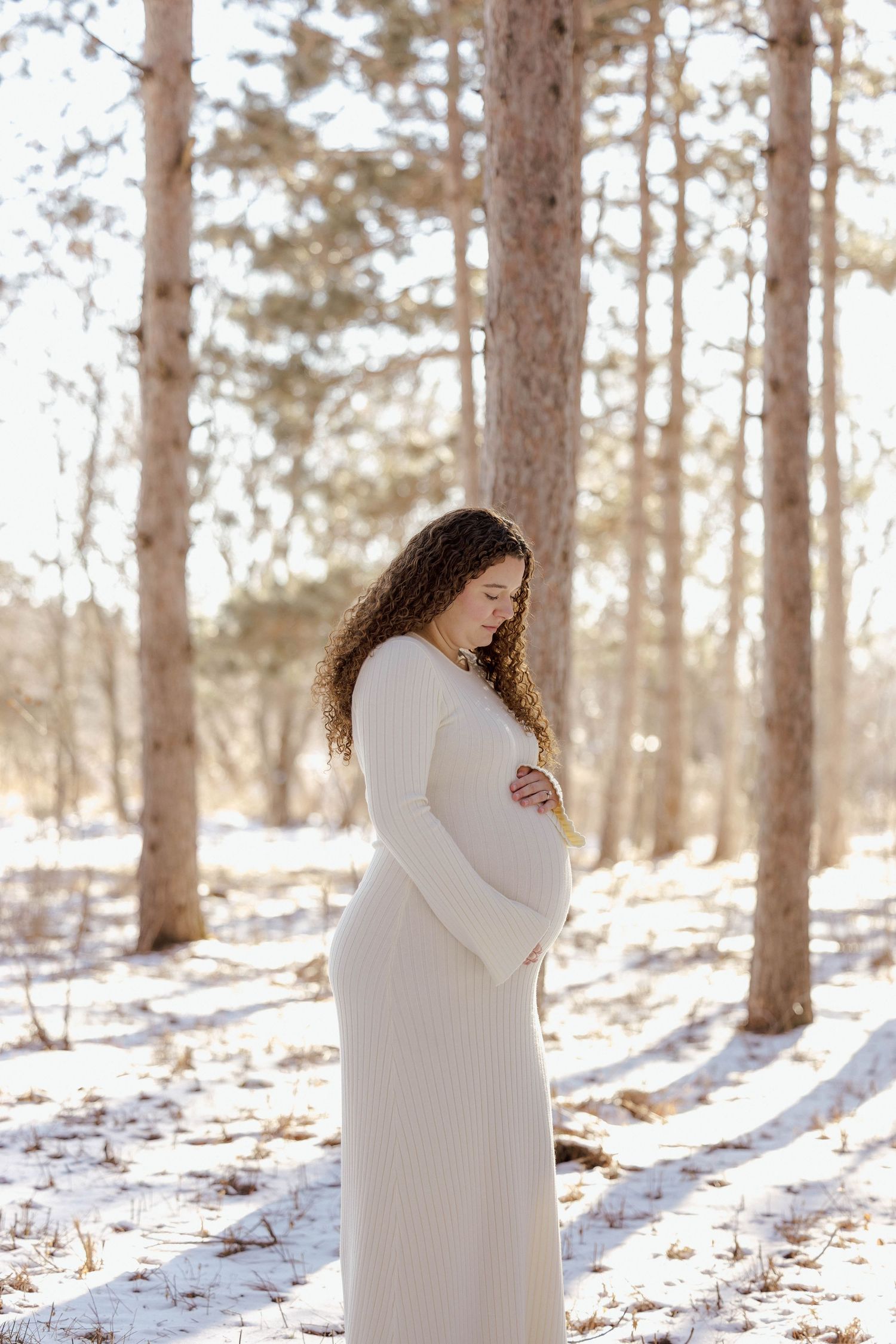 Pregnant woman in cream knit dress cradling belly in snowy winter forest.