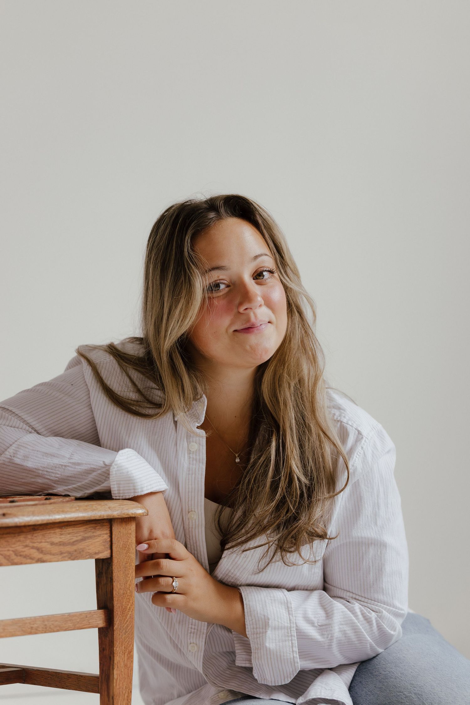 Woman in striped shirt leaning on wooden chair smiling softly indoors.