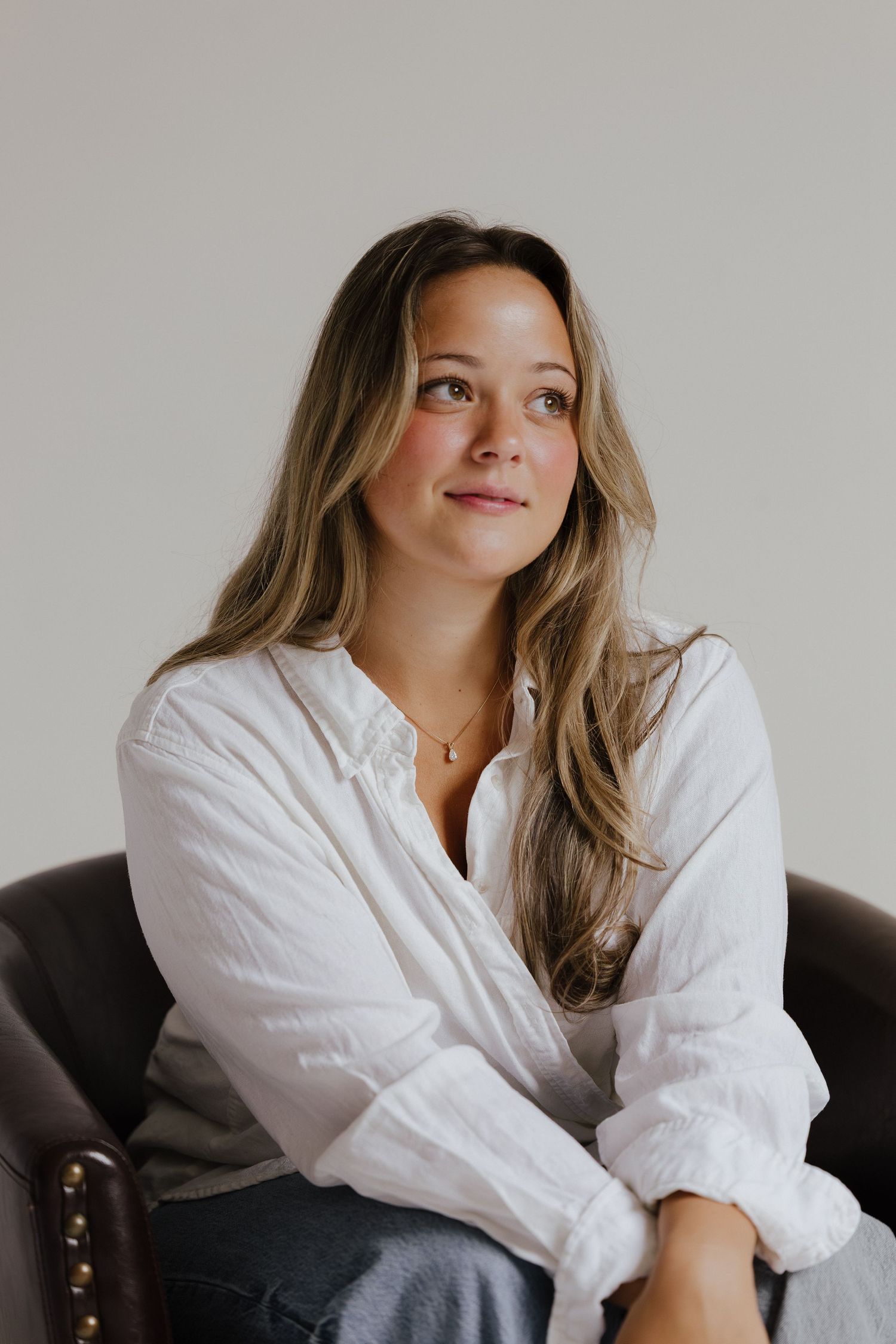 Woman in white linen shirt sitting in leather chair looking up thoughtfully.