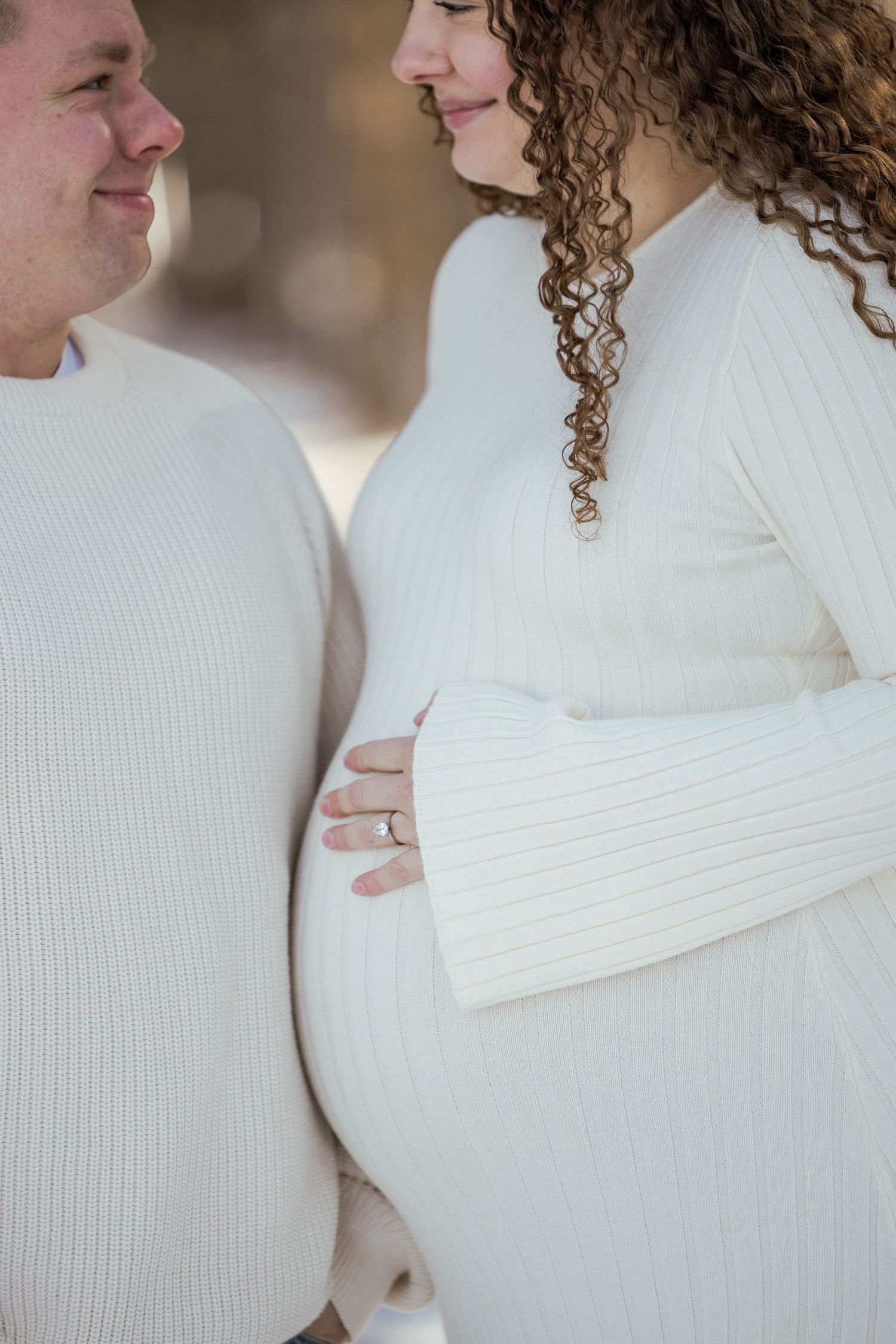 Couple in white sweaters tenderly holding pregnant belly close together outdoors.