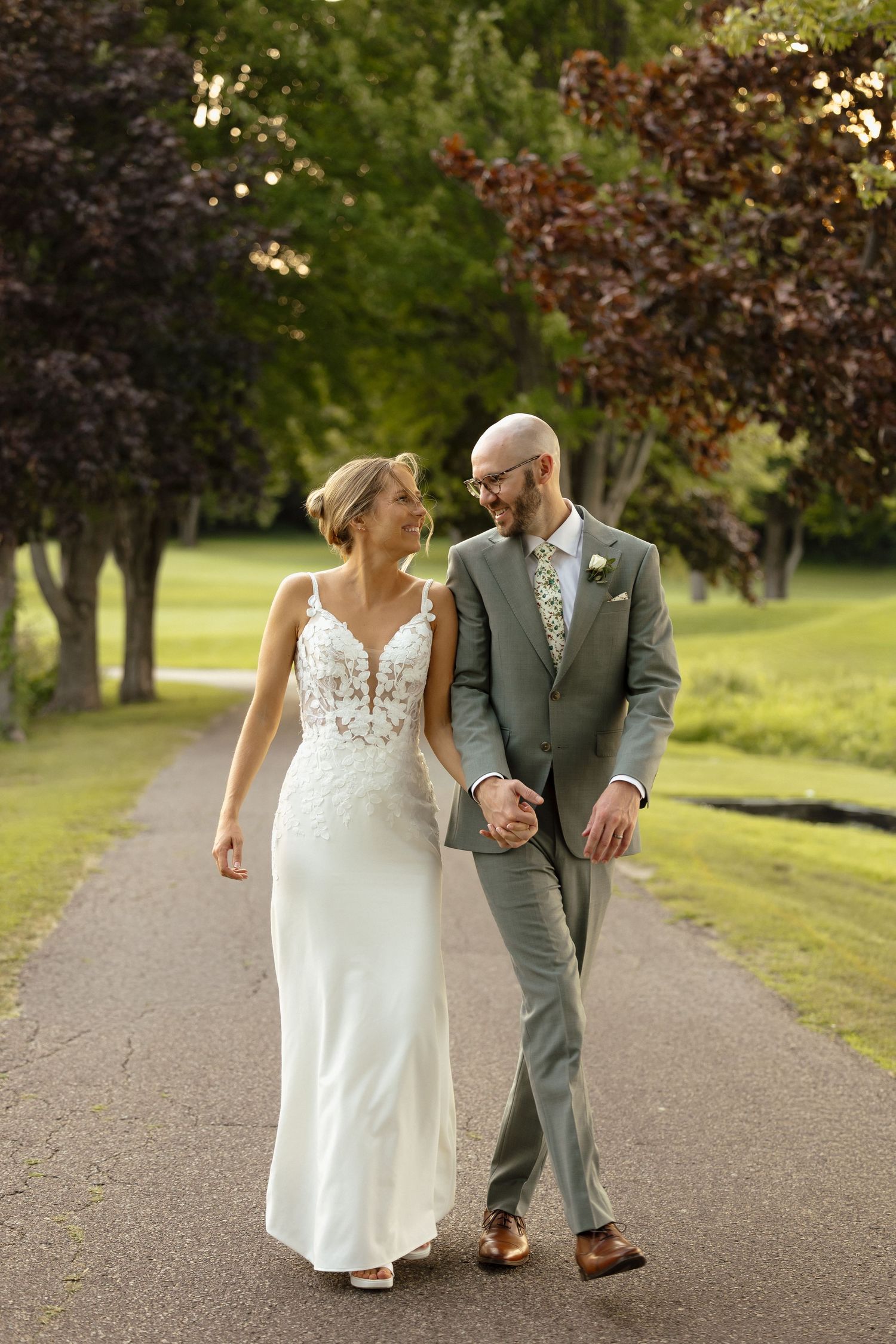 Bride in lace gown and groom in gray suit walk hand in hand along tree-lined path in golden sunlight.