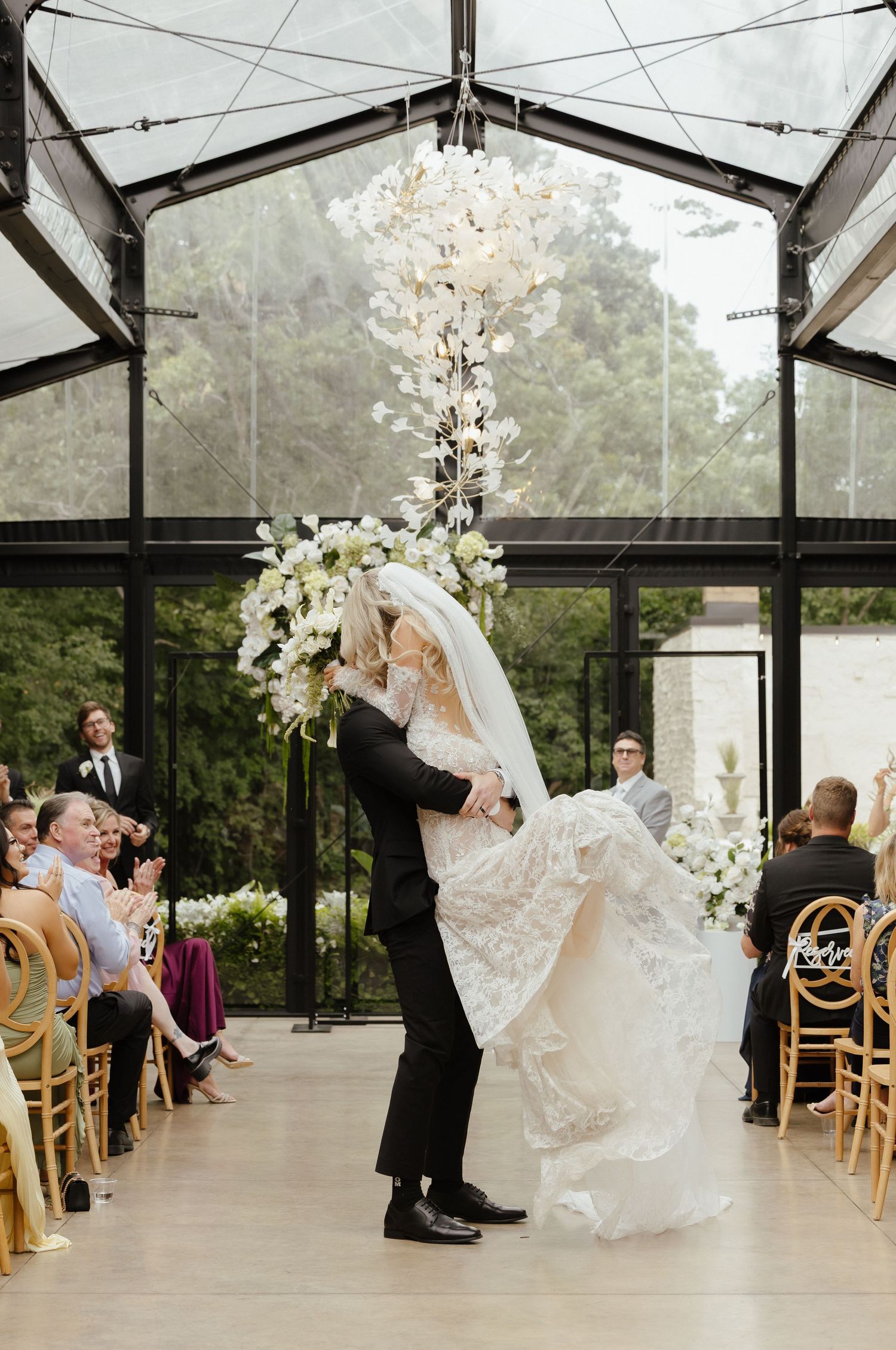 Groom lifts bride at altar in glass greenhouse chapel surrounded by white floral arrangements.