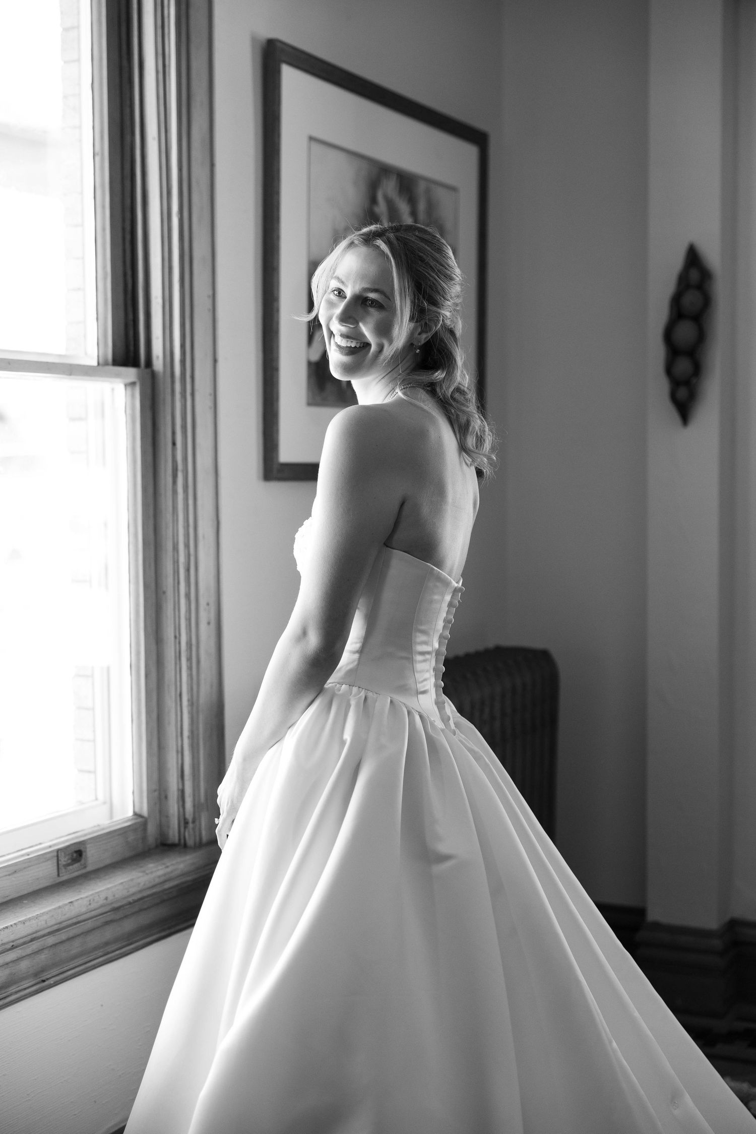 A smiling bride in a strapless ball gown poses by a window in a black and white portrait.