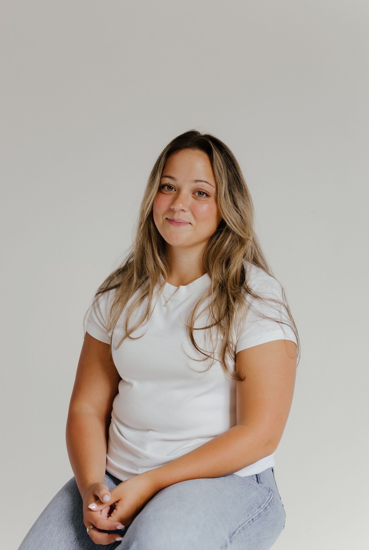Young woman with blonde highlights wearing white fitted tee and jeans sitting and smiling against neutral background.