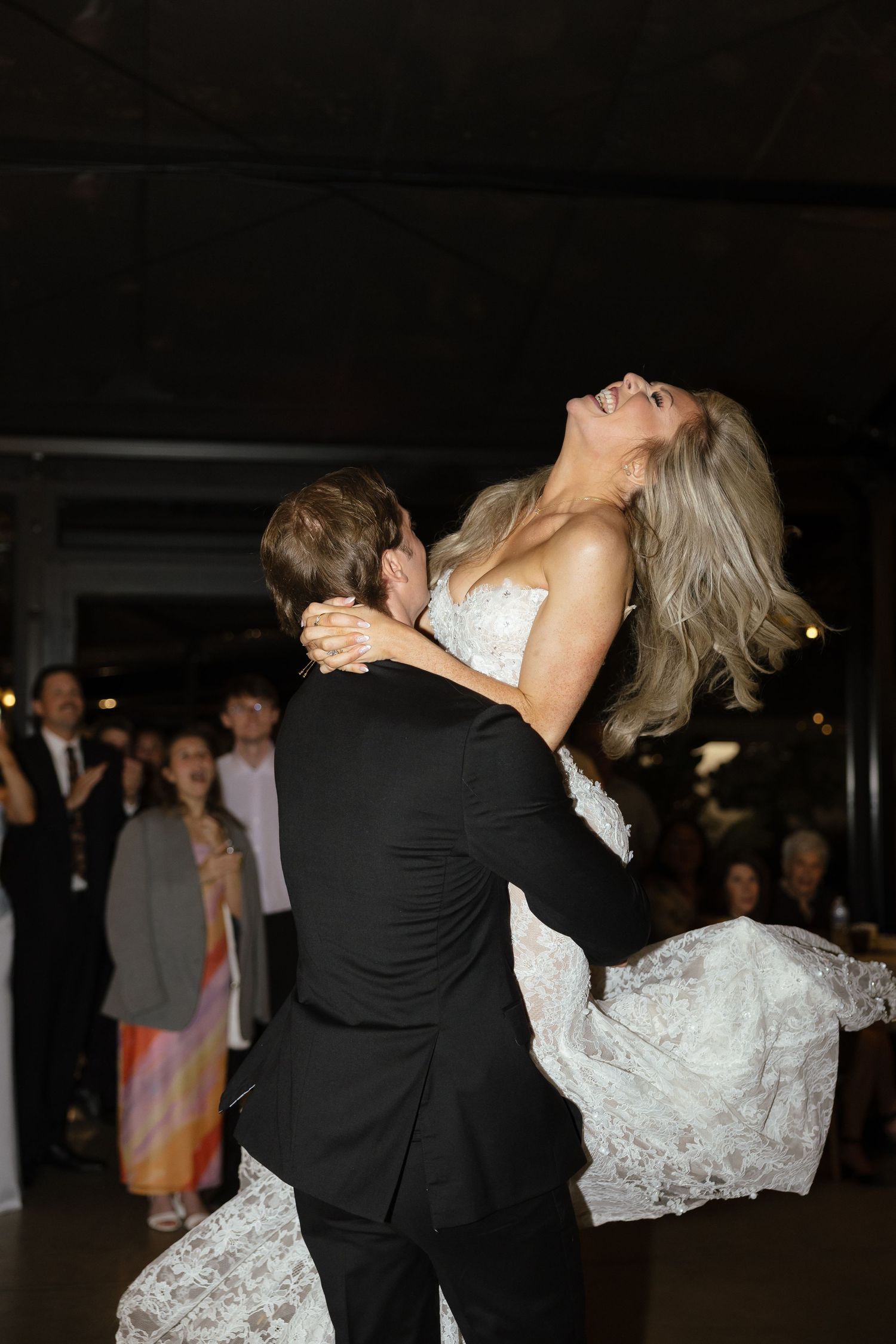 Groom lifts laughing bride during first dance at wedding reception with guests applauding.