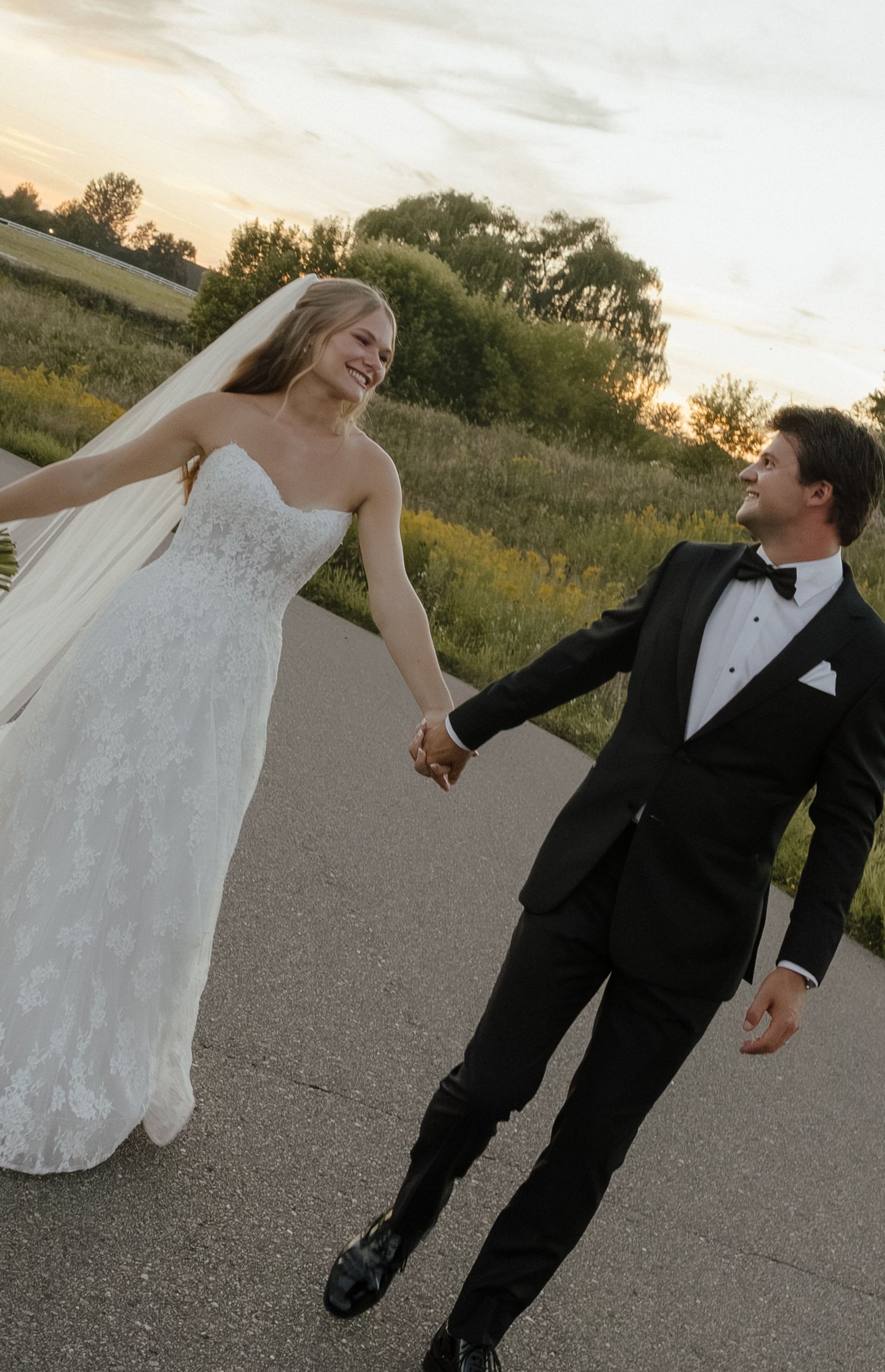 Smiling bride in lace gown holding hands with groom in black tuxedo walking at sunset outdoors.