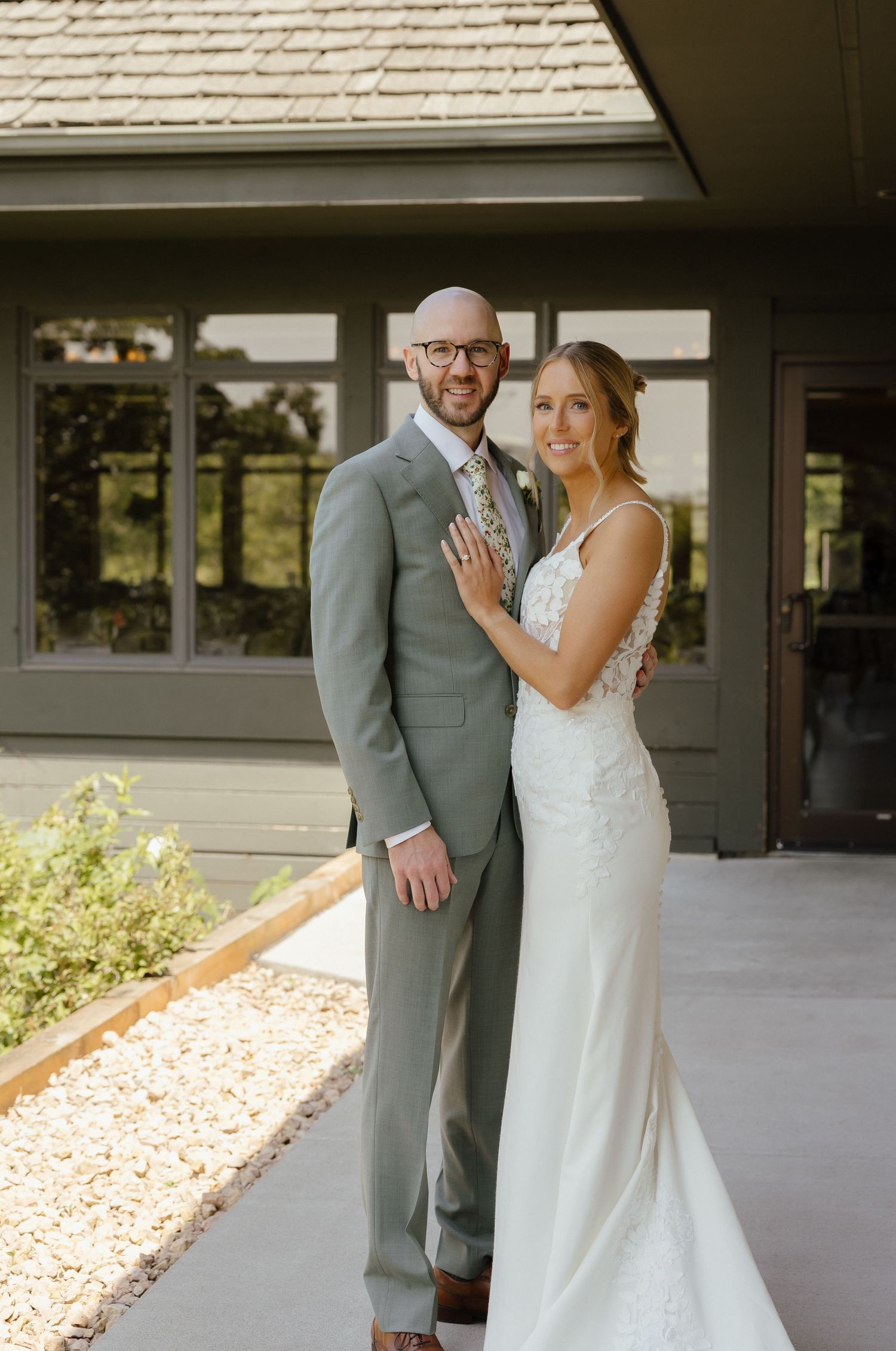 Bride in fitted lace gown and groom in gray suit posing together outside rustic venue.