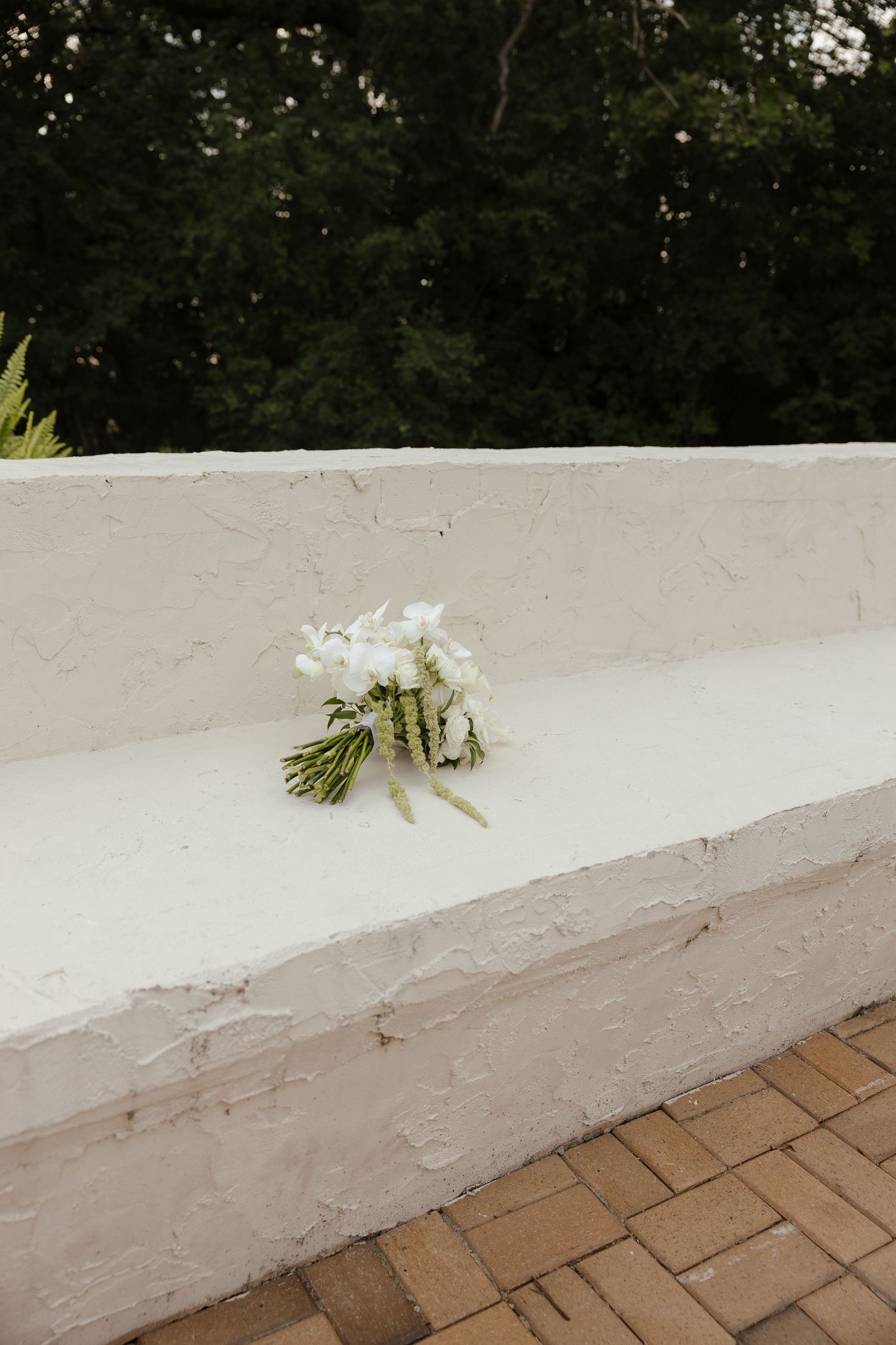 Delicate white floral bridal bouquet resting on a white textured stone bench with brick flooring.